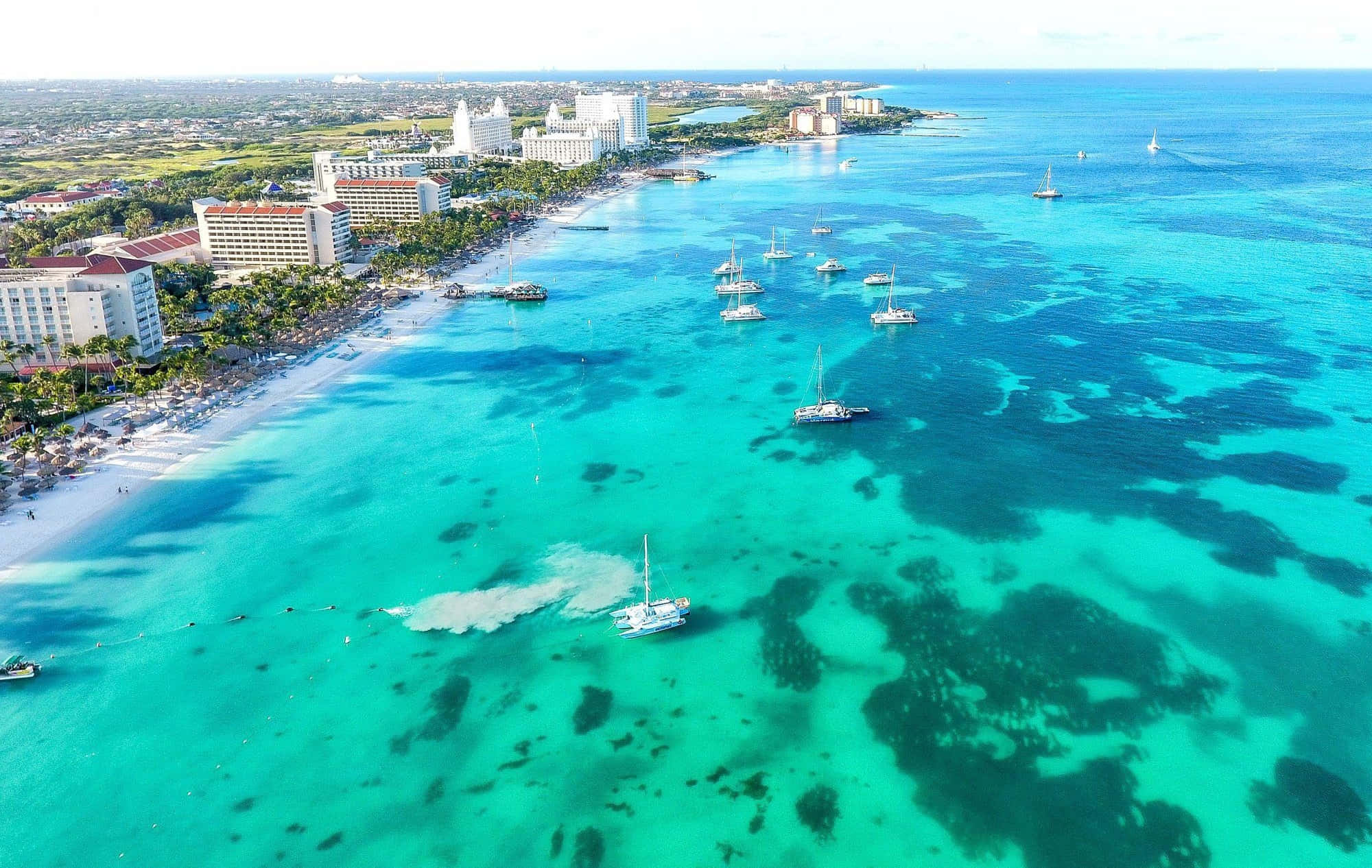 Aruba 桌布 壁紙 — 熱帶海灘的鳥瞰圖，碧綠的海水點綴著帆船，兩旁是白色沙灘度假村，在明亮的日光下。