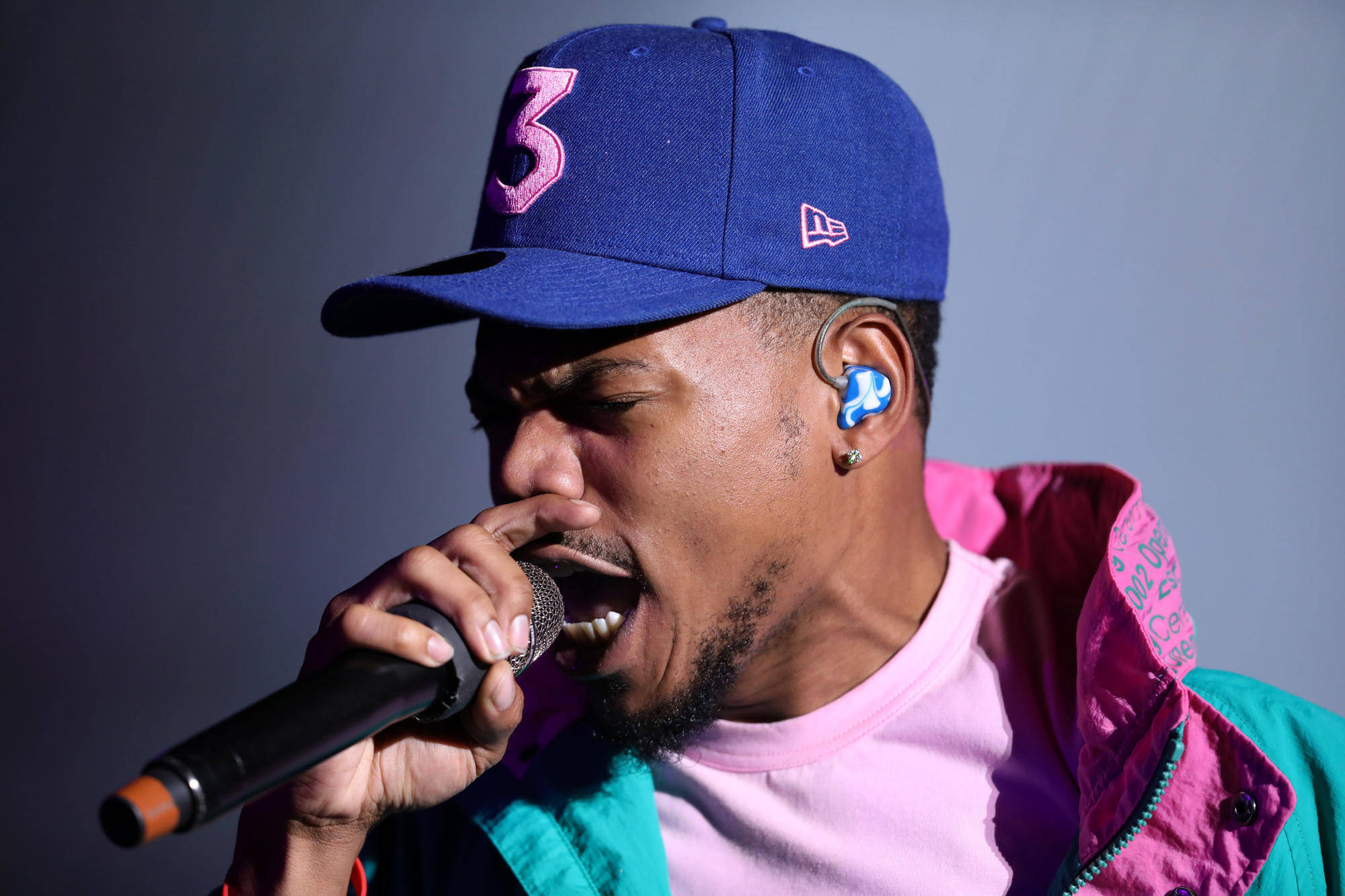 熱情的Chance the Rapper 壁紙 — 一位戴著藍色帽子和彩色外套的男饒舌歌手在舞台燈光下熱情地對著麥克風唱歌。