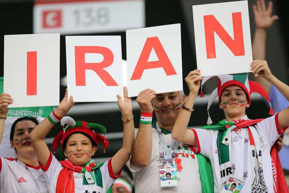 Iran 桌布 壁紙 — 熱情的伊朗球迷自豪地舉起拼寫IRAN的標語牌，身穿國家顏色和臉部彩繪參加體育場活動。