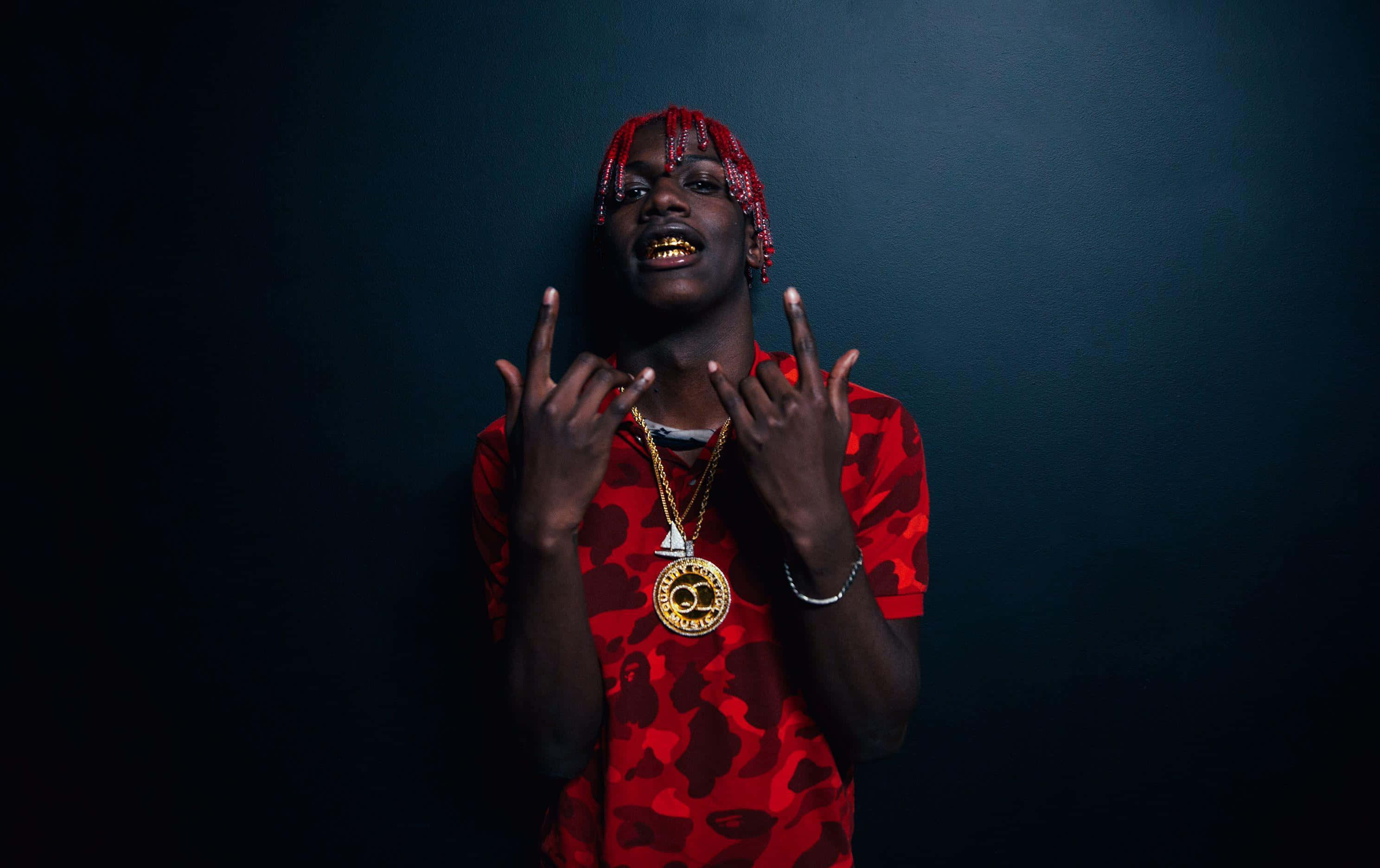 熱情說唱歌手Lil Yachty 壁紙 — 一個有紅色辮子頭髮和金色牙齒的人，穿著紅色迷彩襯衫和金色鏈條，在深藍色背景前做手勢。