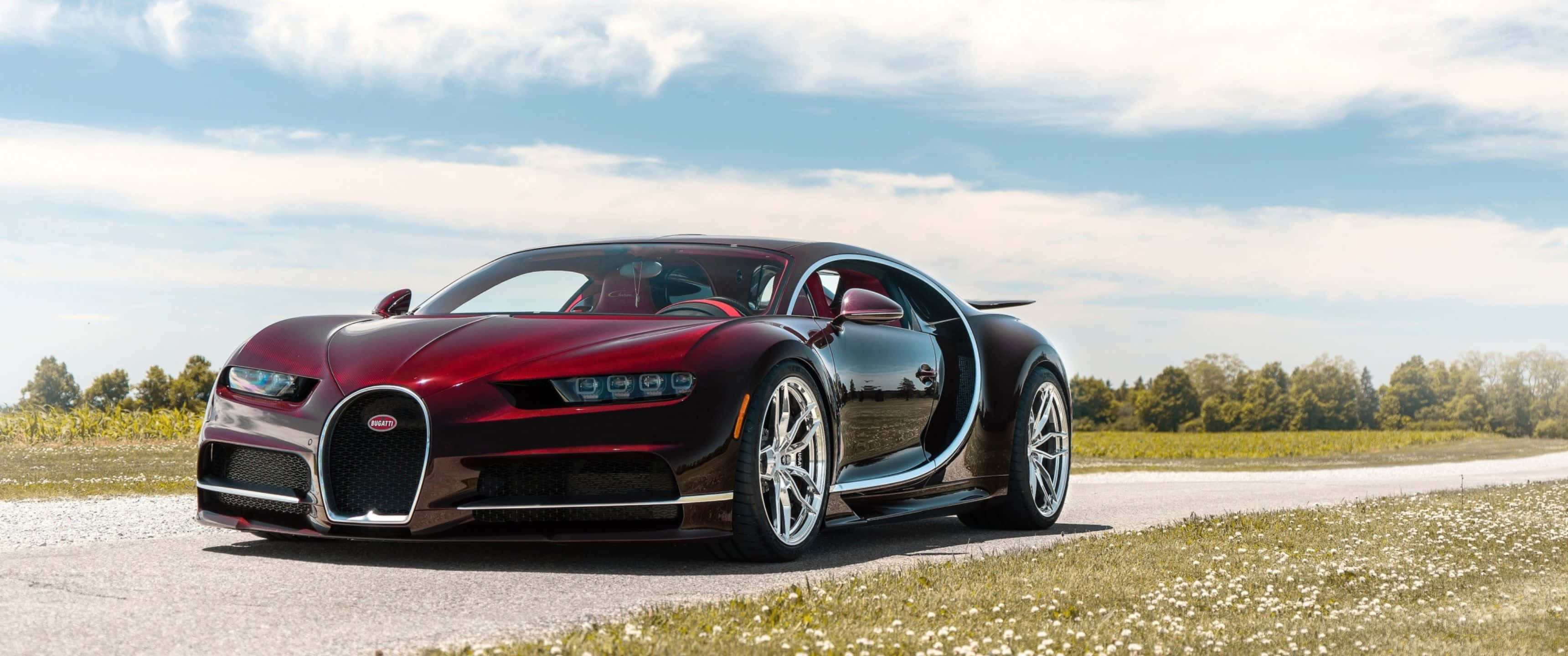 熱門車款！欣賞這輛光亮如新的 Bugatti Chiron Sport 壁紙 — 一輛光澤的栗色Bugatti Chiron打開剪刀門，停在碎石路上，旁邊是野花田，天空部分多雲。