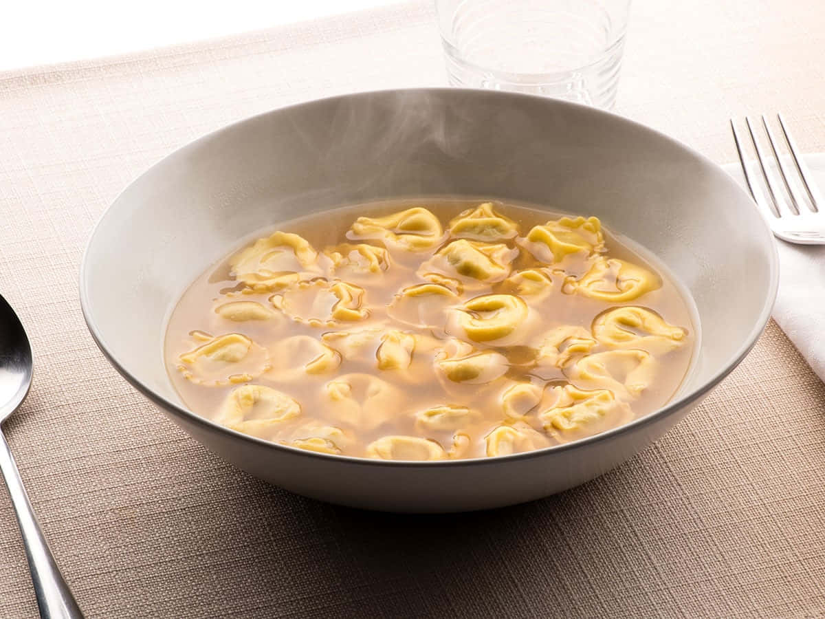 熱騰騰的義式餃子湯 壁紙 — 一碗金黃色意大利雲吞在忌廉湯中冒著蒸氣，放在有紋理的米色枱布上，旁邊有餐具。