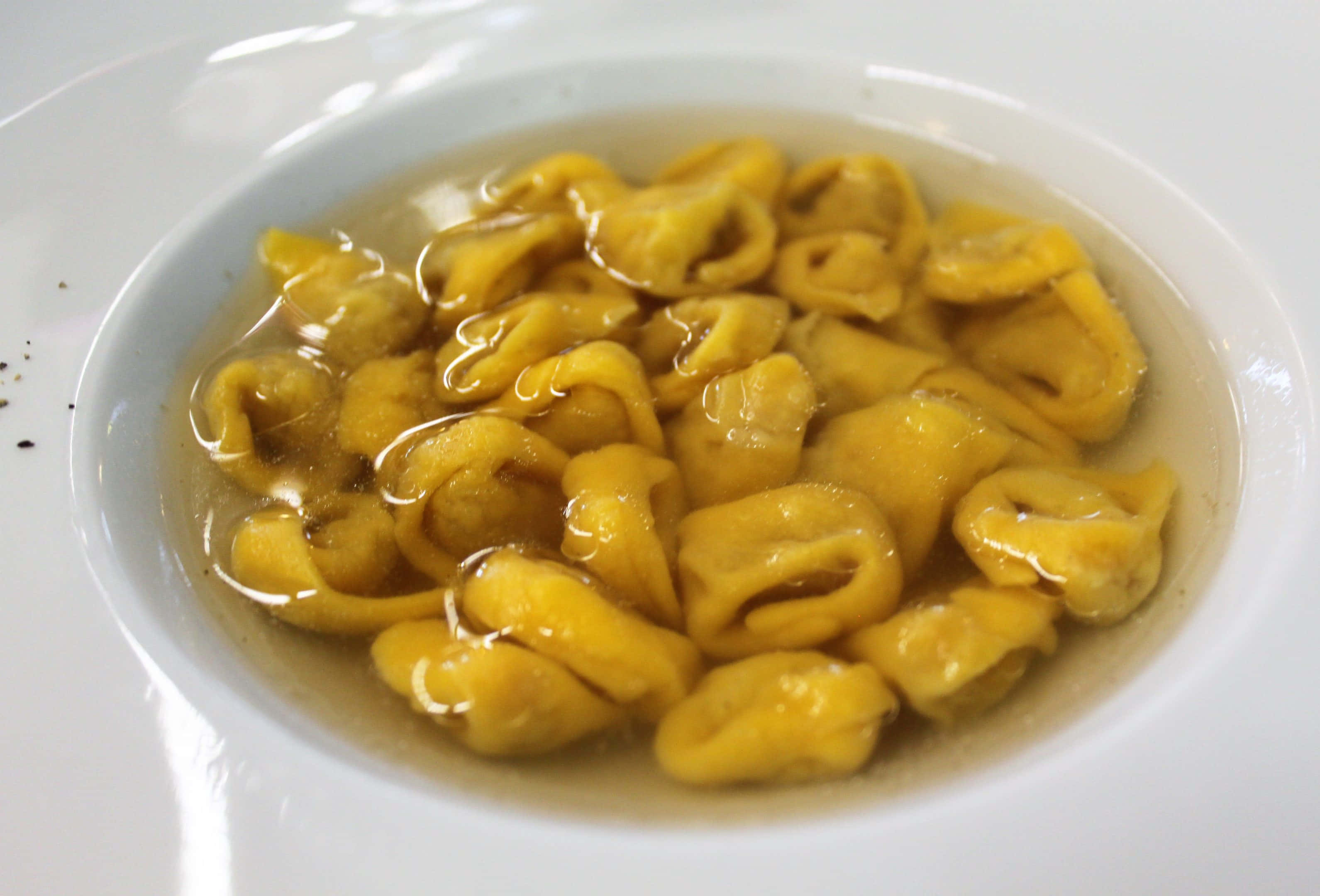 熱騰騰的義式餃子湯 壁紙 — 白色碗中清澈湯汁裡漂浮著金色意大利餃子的特寫，碗邊有少許黑胡椒碎。