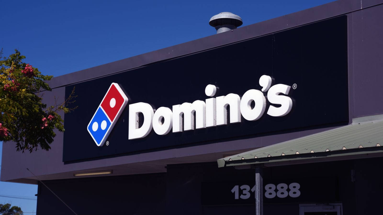 熱鬧多米諾披薩門市夜景 壁紙 — 晴朗的藍天襯托著 Domino's Pizza 店面，白色粗體字和紅藍標誌在清晰日光下格外醒目。