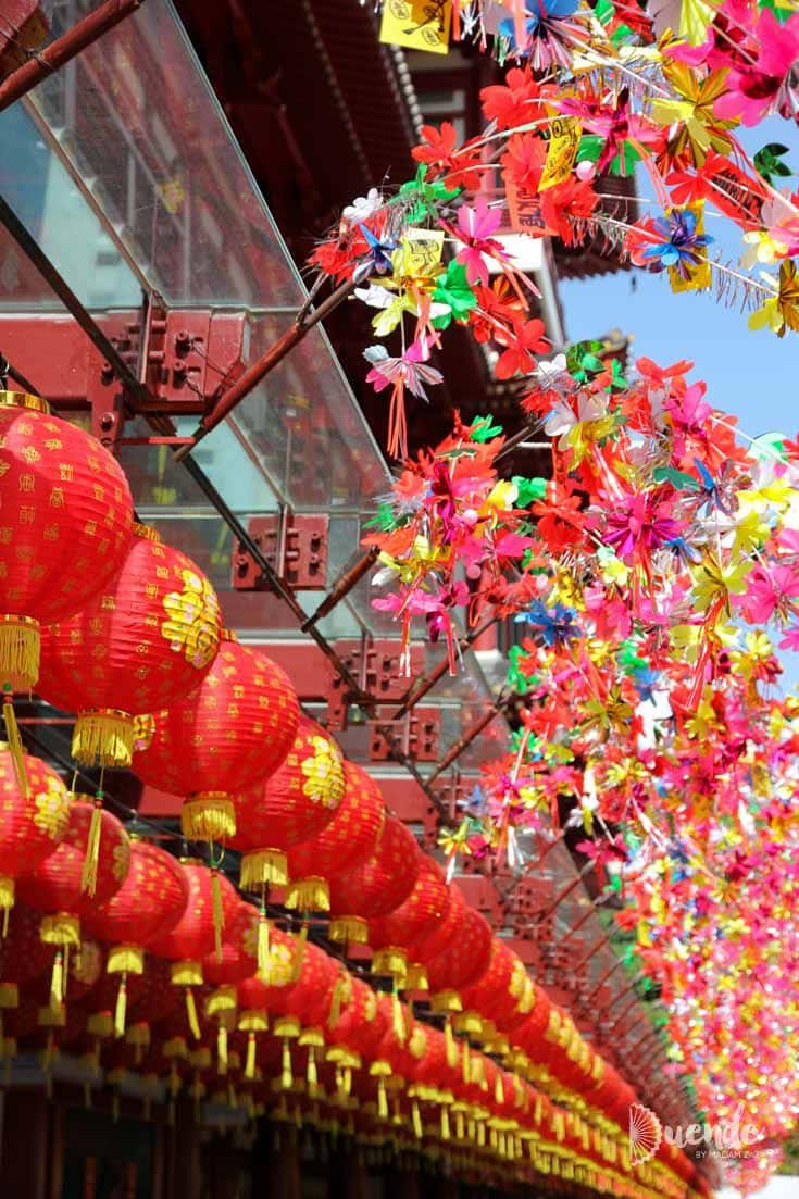 熱鬧的中國新年慶祝活動 壁紙 — 鮮豔的紅色燈籠和色彩繽紛的紙花在明亮的藍天下，喜慶地掛在傳統中式寺廟屋頂上。