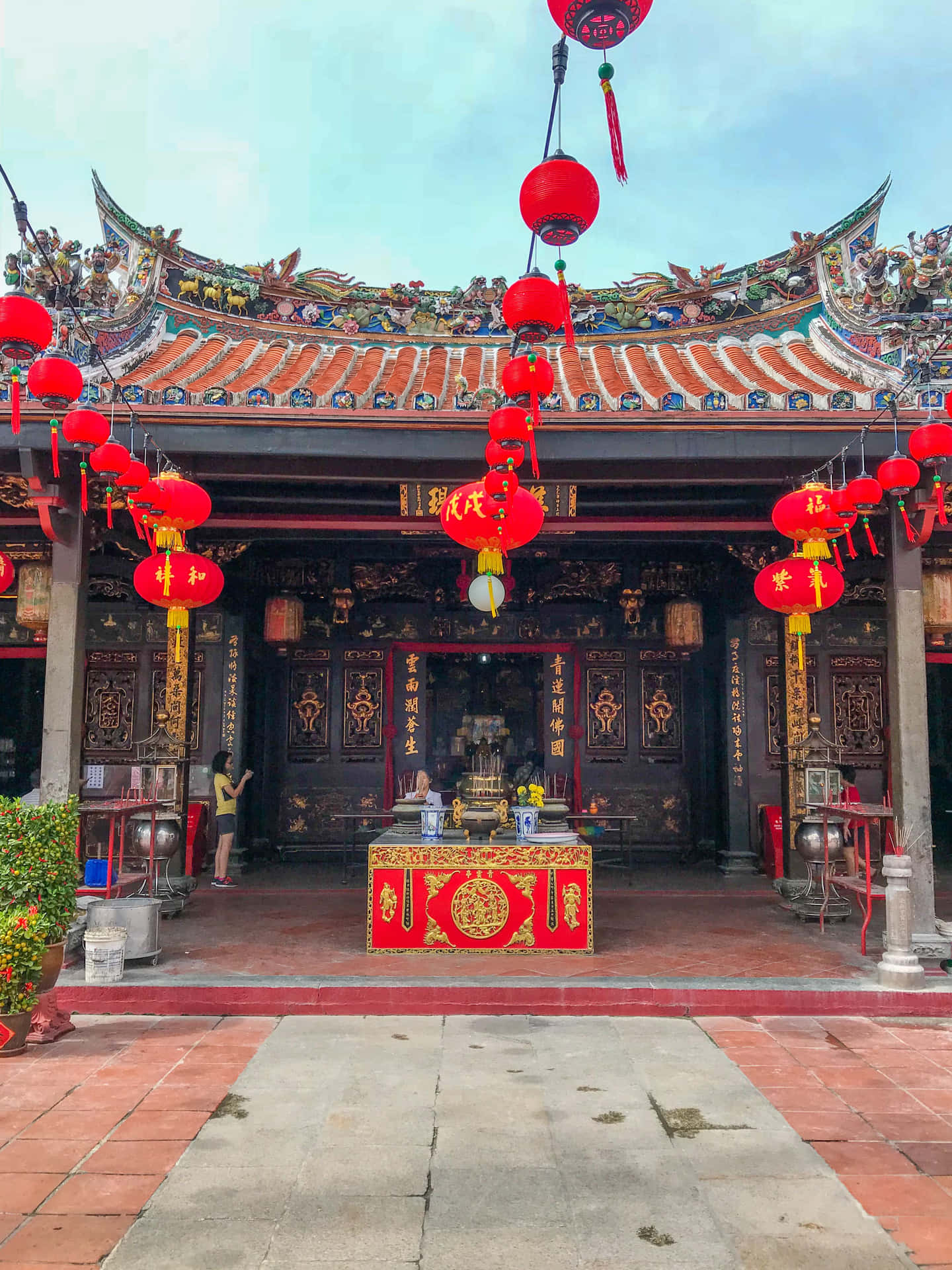 熱鬧的中國新年慶祝活動 壁紙 — 傳統中國寺廟，擁有華麗的瓦片屋頂和紅色燈籠，懸掛在紅色和金色的祭壇上方，天空晴朗。