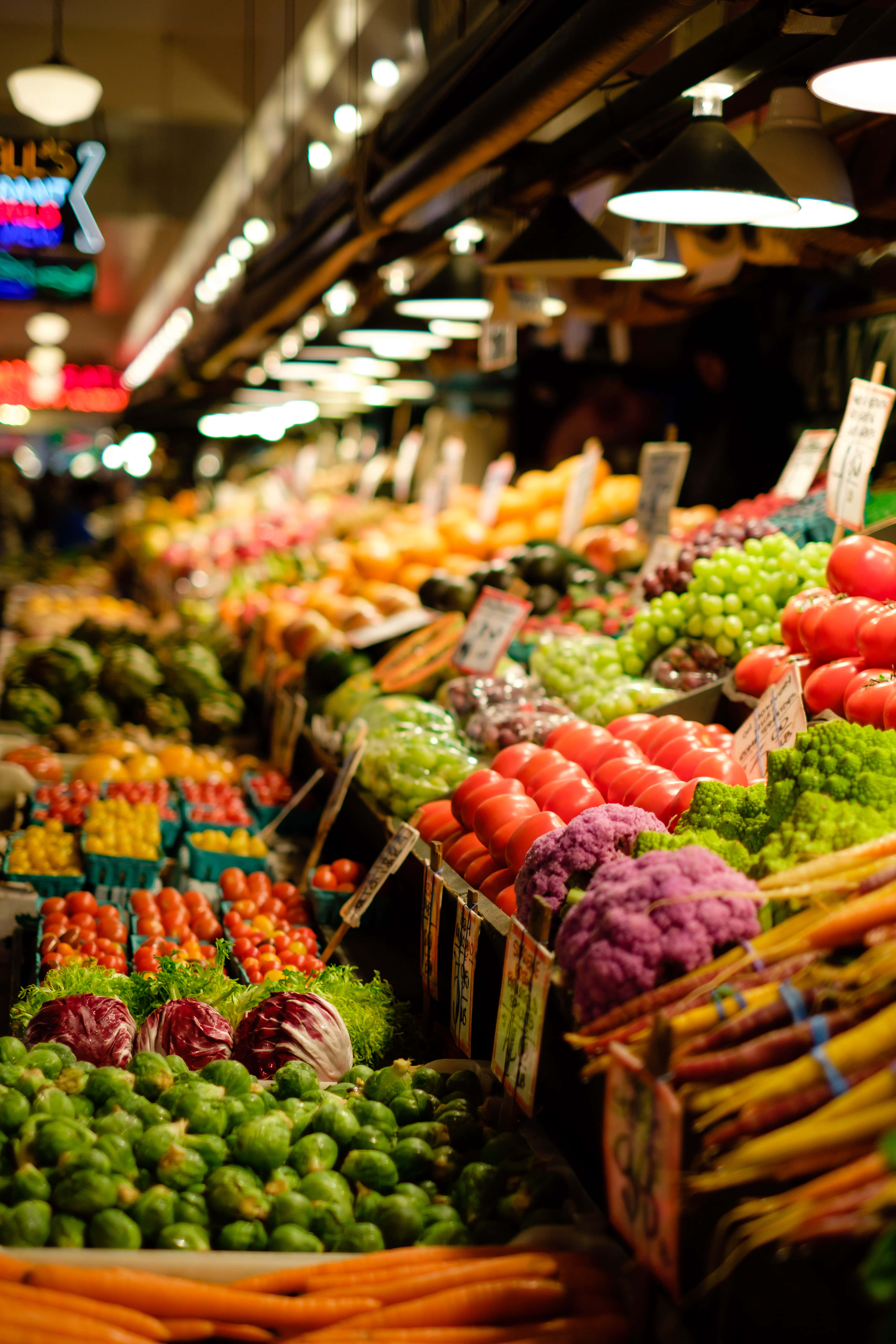 Grocery Store 桌布 壁紙 — 熱鬧的室內市場中，色彩繽紛的農產品攤位在溫暖的吊燈下閃耀。