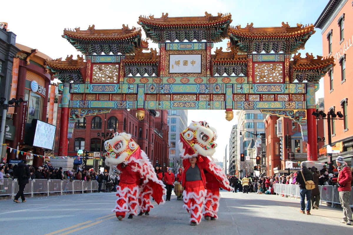 熱鬧的春節慶祝活動 壁紙 — 充滿活力的紅色舞獅在繁華的城市街頭節日中，於傳統中式牌坊下表演。