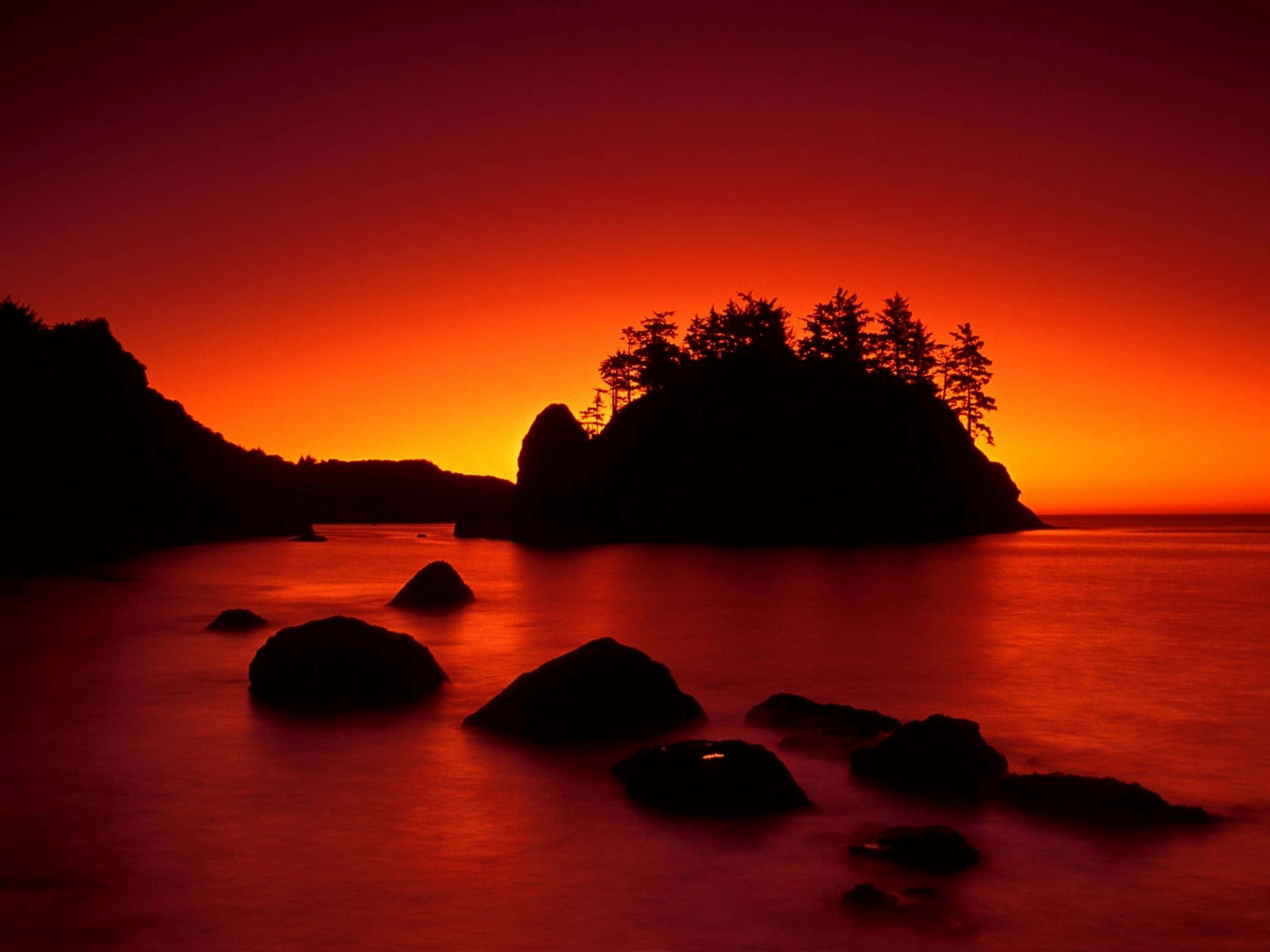 Red Sunset 桌布 壁紙 — 熾熱的夕陽將天空和水面染成深紅和橙色，輪廓分明的岩石和樹木在發光的地平線上顯得格外突出。