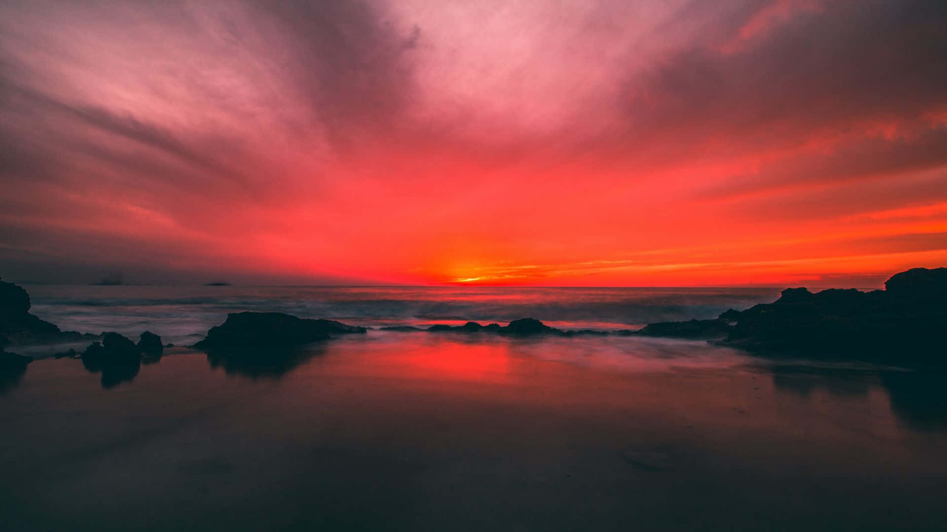 海灘日落 桌布 壁紙 — 熾熱的夕陽將天空和海洋染成鮮豔的紅色和橙色，海浪輕輕拍打著黑暗的岩石輪廓。