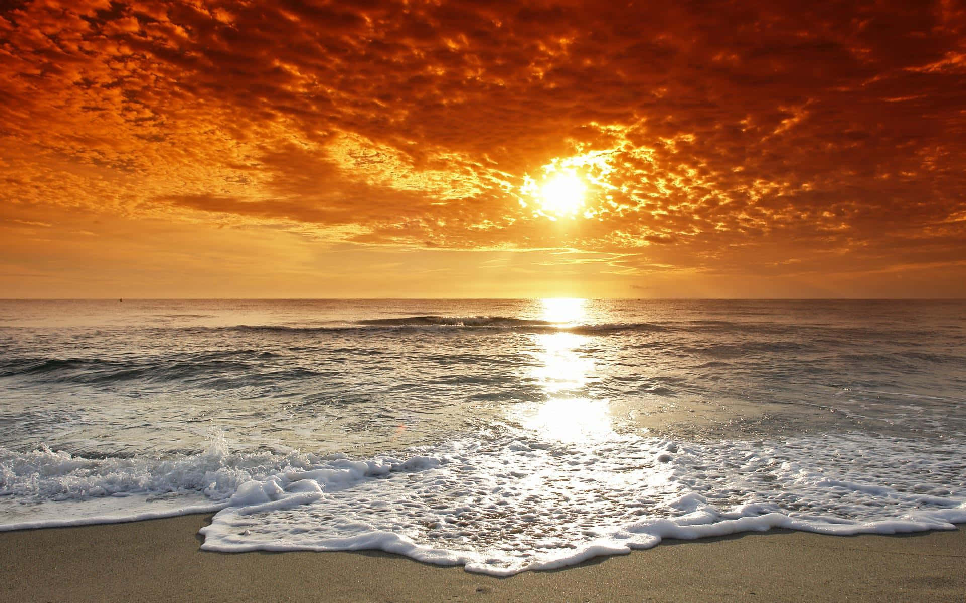 海灘日落 桌布 壁紙 — 熾熱的夕陽將天空染成橙色，海浪拍打沙灘，太陽在水面上反射。