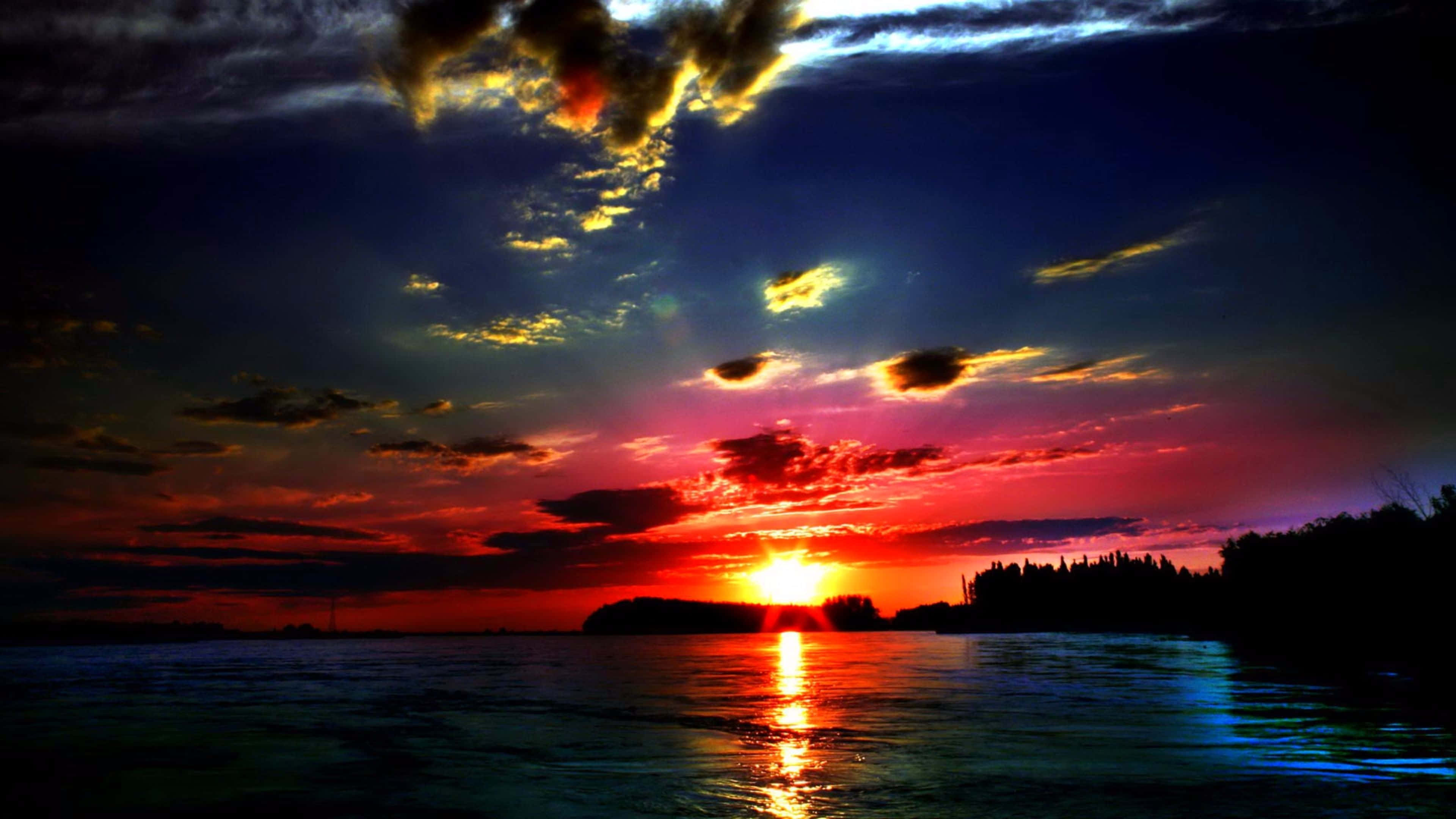 Sunset View 桌布 壁紙 — 熾熱的夕陽將天空染成紅色、紫色和金色，太陽沉入遠處的樹線之下，倒映在平靜的水面上。