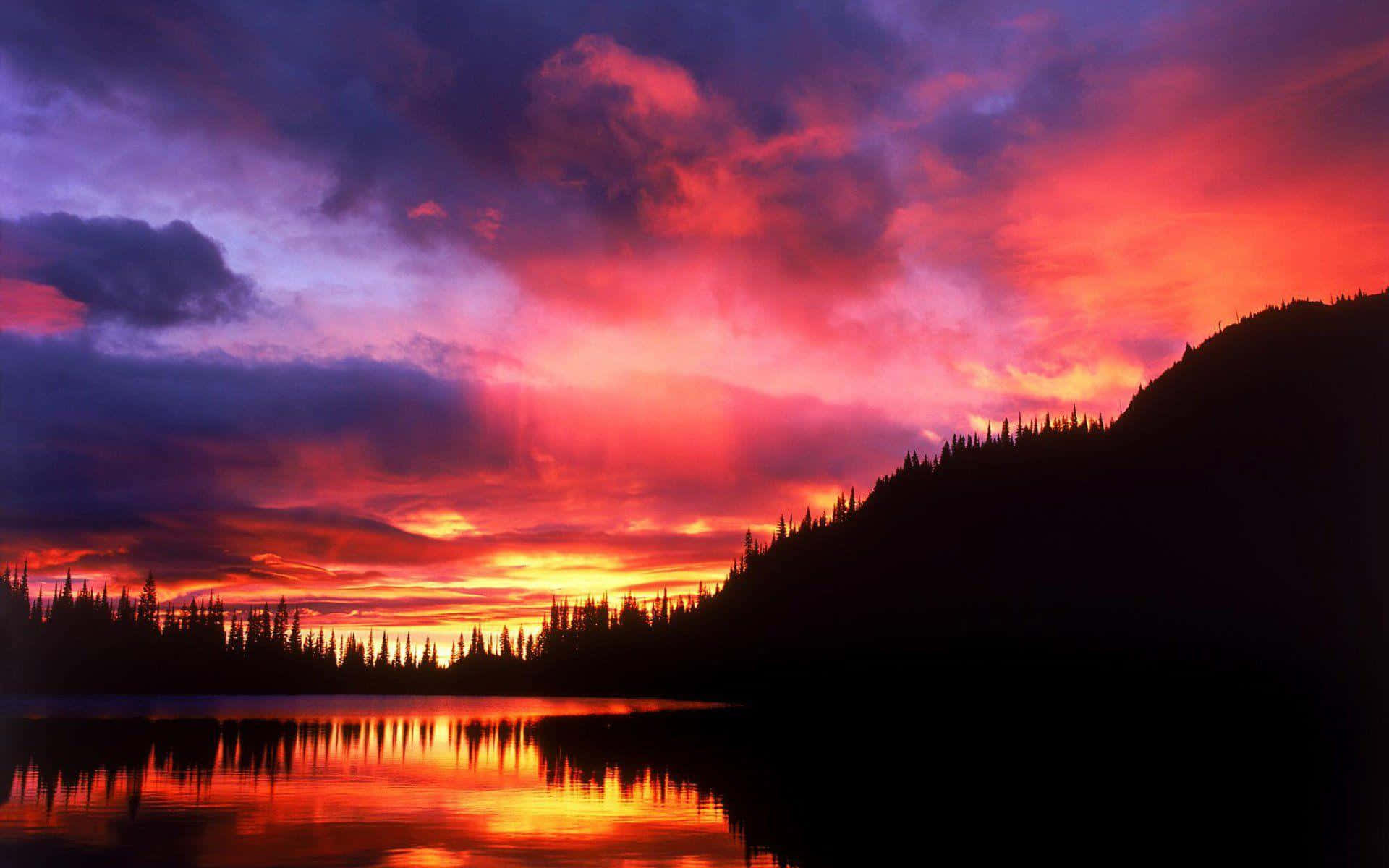 Sunset Landscape 桌布 壁紙 — 熾熱的夕陽將天空染成鮮豔的紅色、紫色和橙色，完美地倒映在平靜的湖面上，周圍是黑暗的森林山丘。