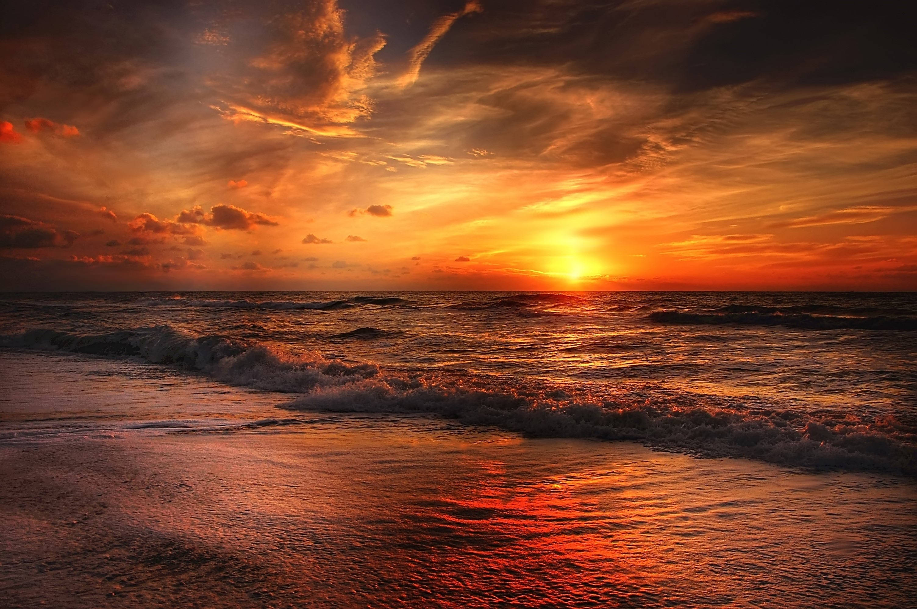 海灘日落 桌布 壁紙 — 熾熱的夕陽將海洋和天空染成鮮豔的橙色和紅色，海浪拍打著濕漉漉的沙灘。