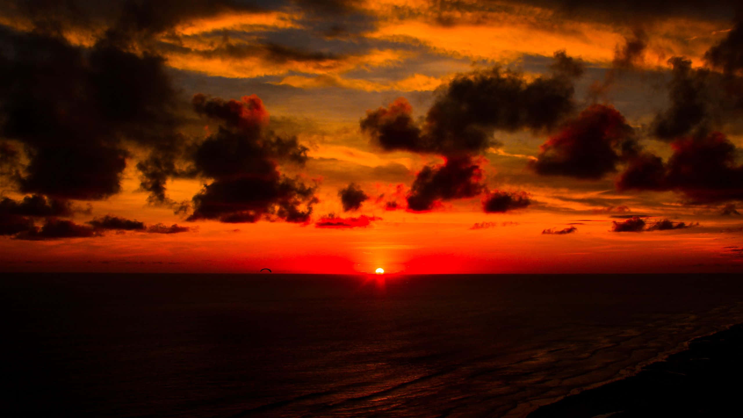 Red Sunset 桌布 壁紙 — 熾熱的夕陽用熔化的橙色和紅色描繪海洋地平線，暗雲在發光的天空中形成剪影。
