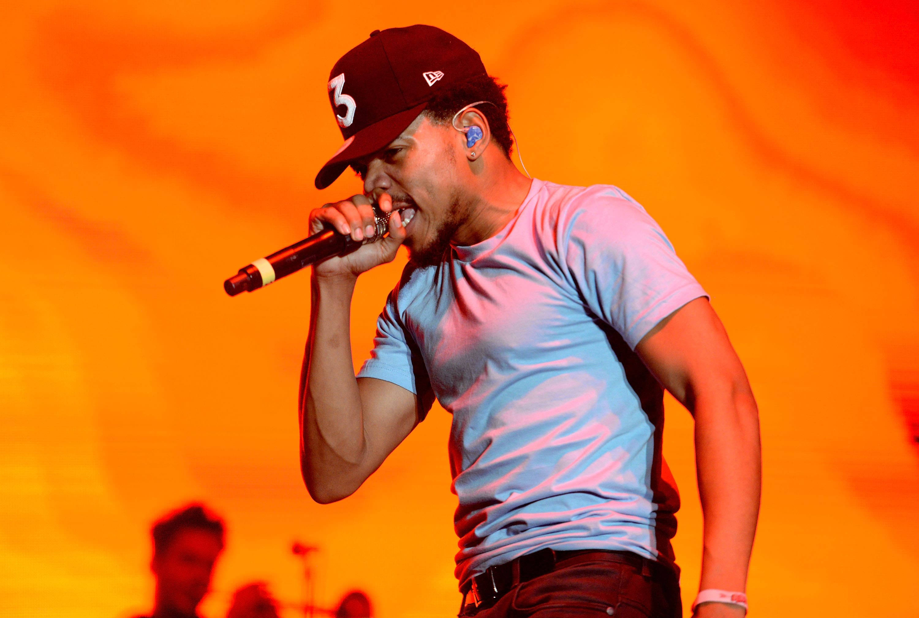 燃燒的Chance the Rapper 壁紙 — 一位戴著帽子的男饒舌歌手在充滿活力的橙色舞台背景下，熱情地對著麥克風表演。