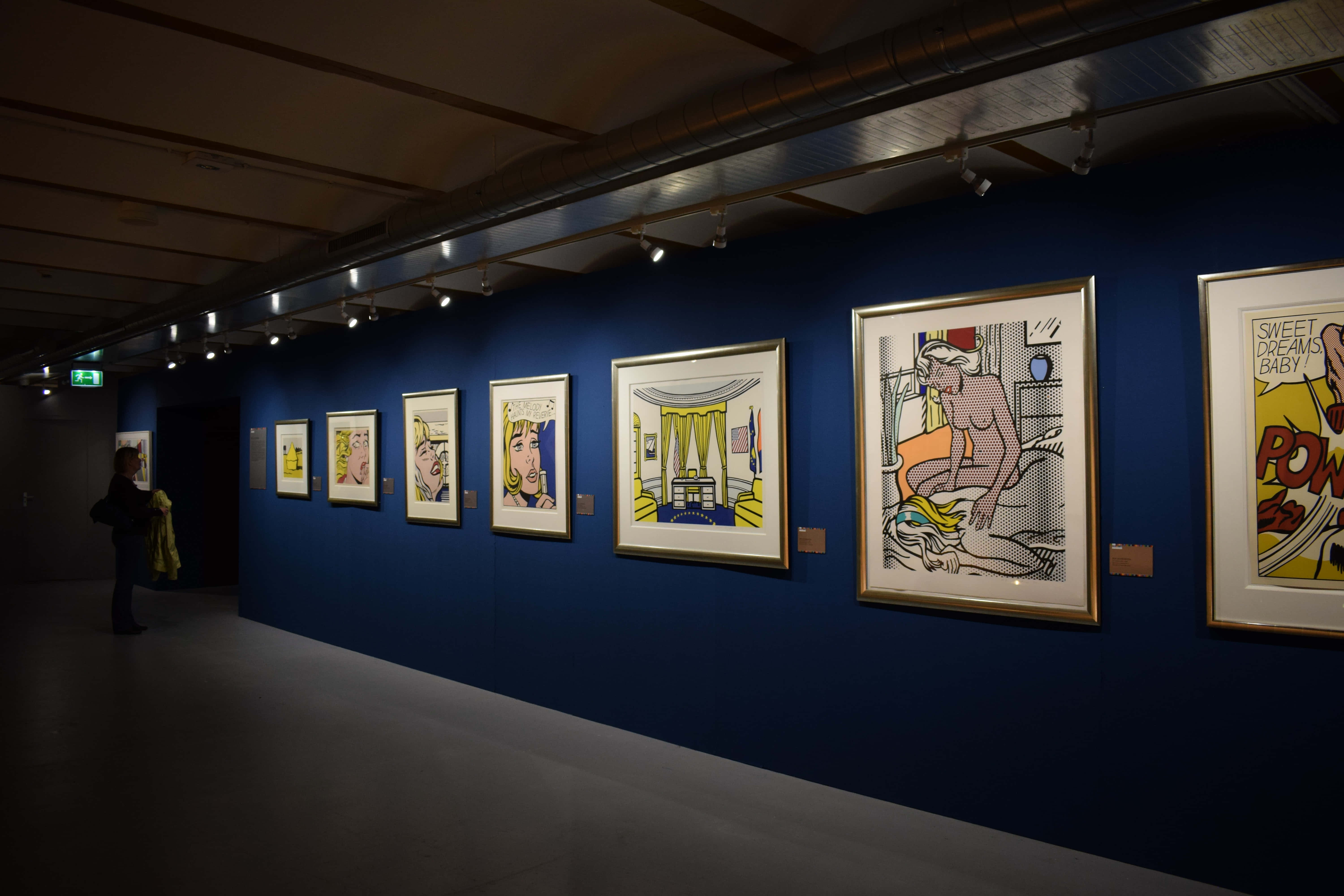 Exhibition 桌布 壁紙 — 燈光昏暗的藝術畫廊，深藍色牆壁上展示著聚光燈下的裱框流行藝術印刷品，遠處站著一位訪客。
