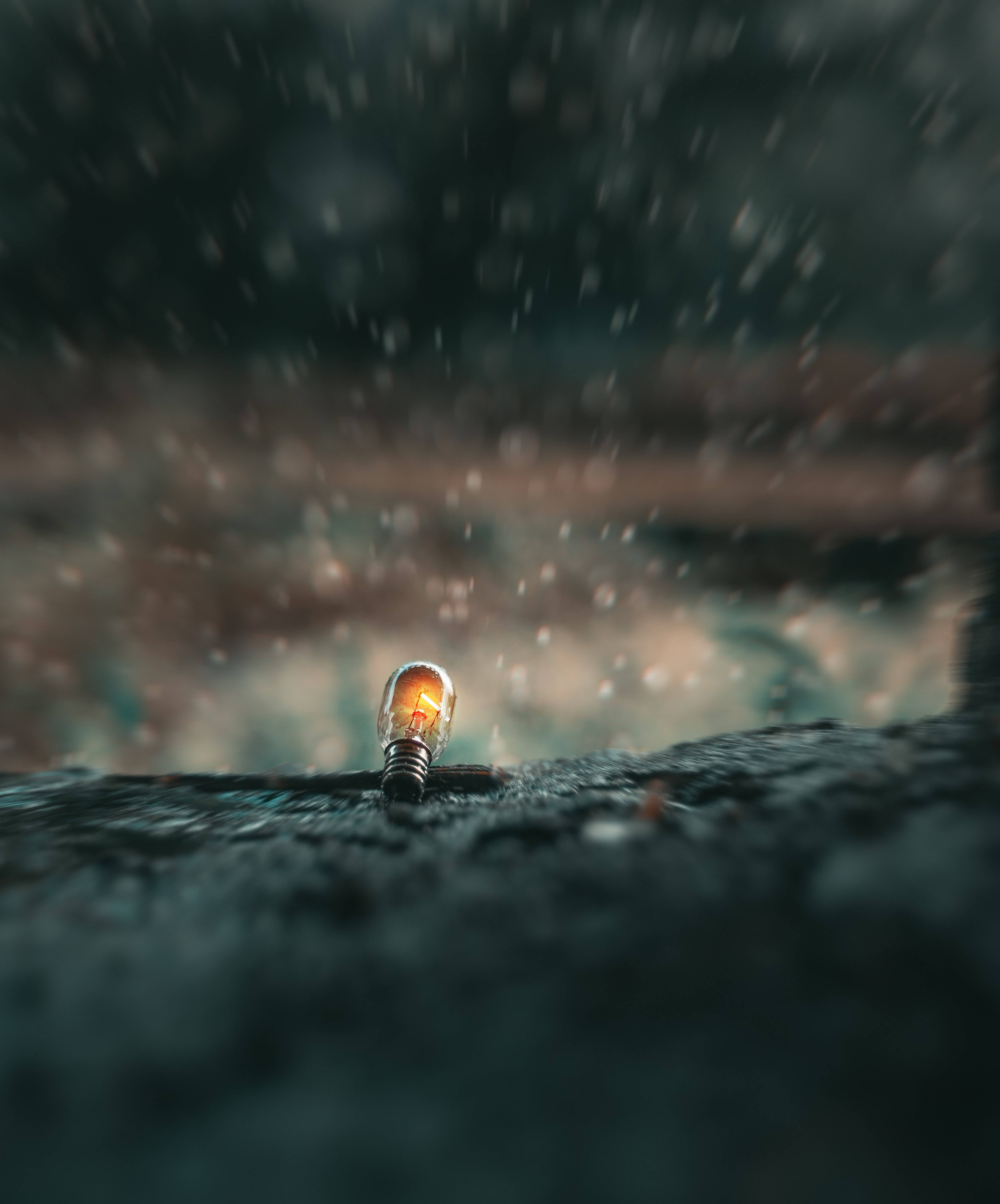 燈泡聚焦深色背景 壁紙 — 一盞發光的燈泡在潮濕的表面上挑釁地閃爍，雨滴落在周圍，營造出陰鬱、充滿氛圍的場景。