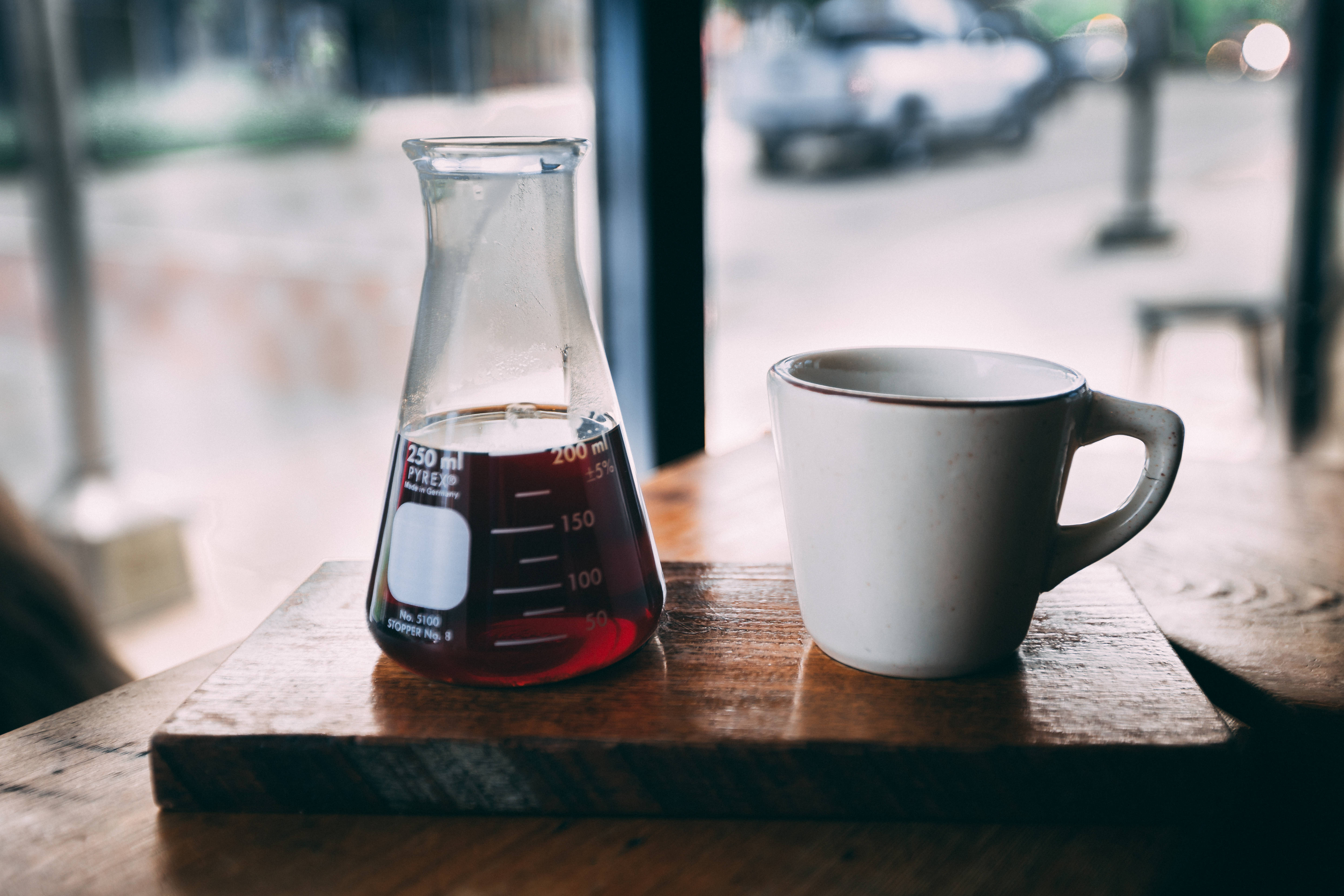 燒杯與咖啡杯 壁紙 — 一個裝滿黑咖啡的玻璃燒杯放在窗邊的木托盤上，旁邊有一個白色馬克杯，窗外是模糊的街景。