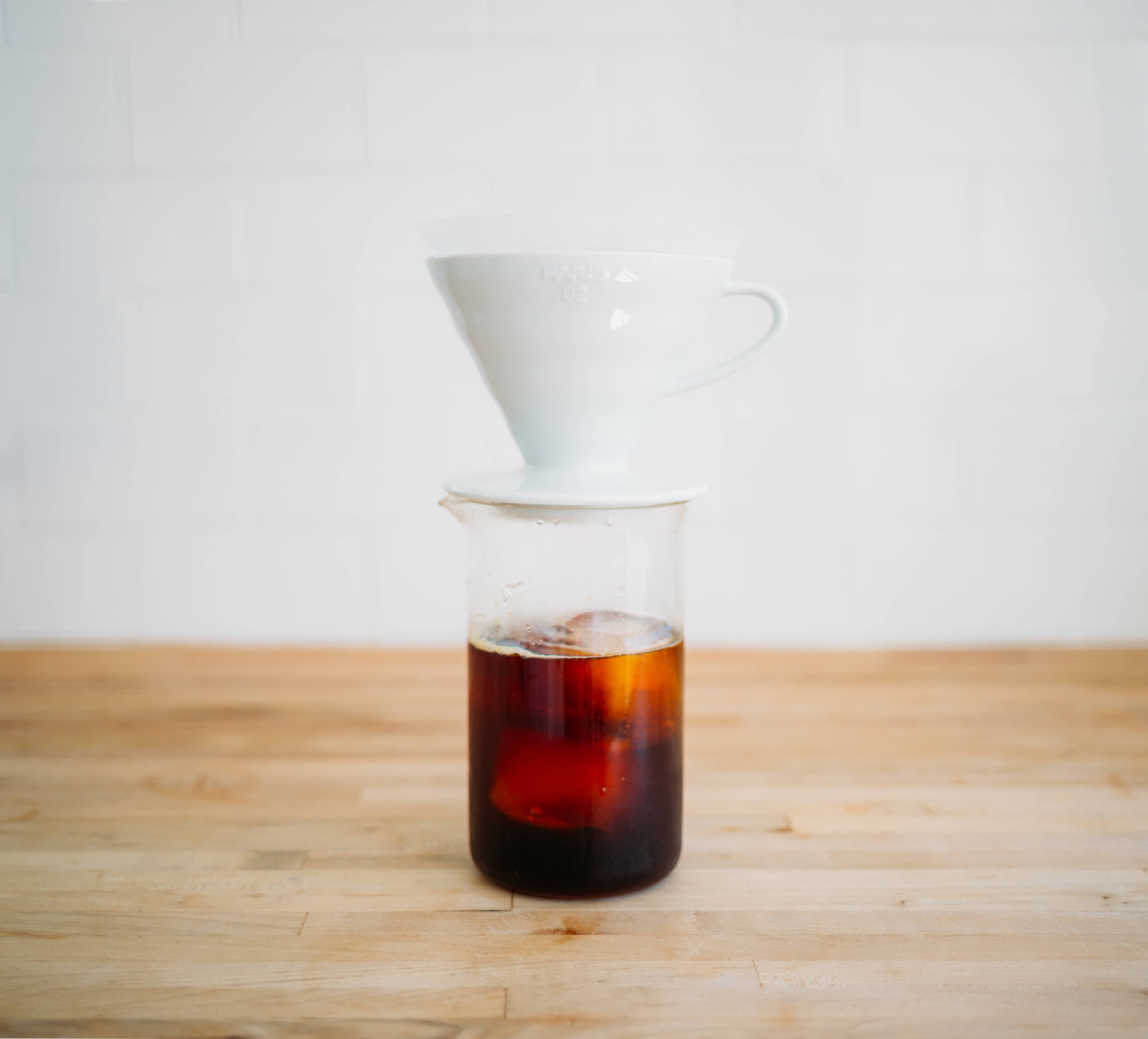 燒杯配冰咖啡與白杯 壁紙 — 一個白色陶瓷手沖咖啡濾杯放在裝滿濃黑咖啡的玻璃咖啡壺上，置於淺色木枱面。