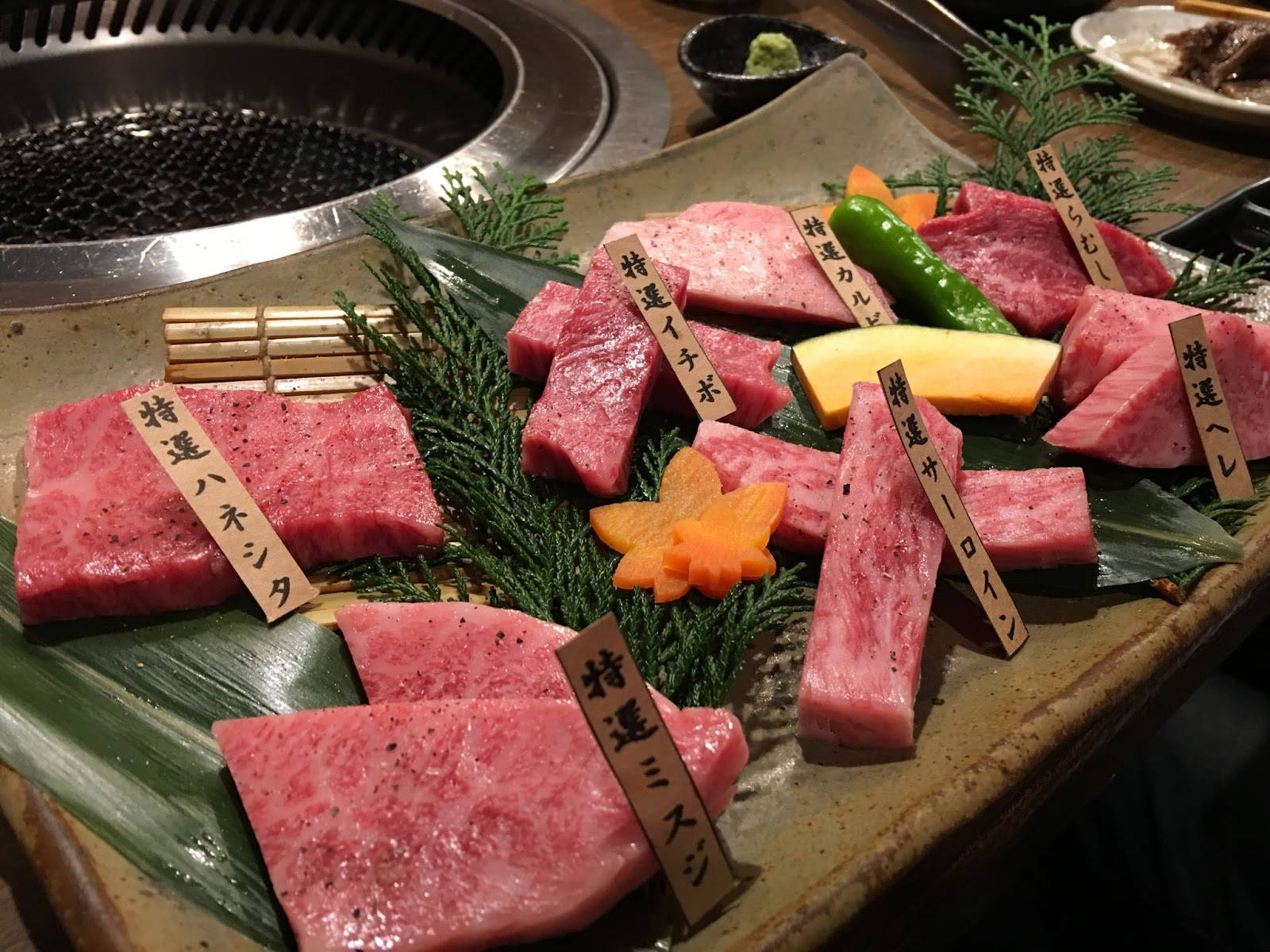 一盤優質和牛肉片，附有木牌和配菜，放在熱烤架旁。