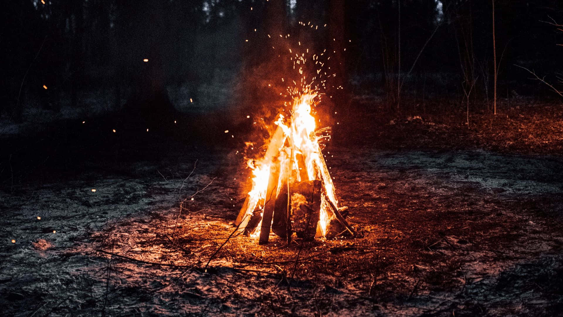 營地中的營火 壁紙 — 一個咆哮的營火在夜晚將溫暖的橙色光芒和飛舞的火花投射到黑暗的森林地面上。