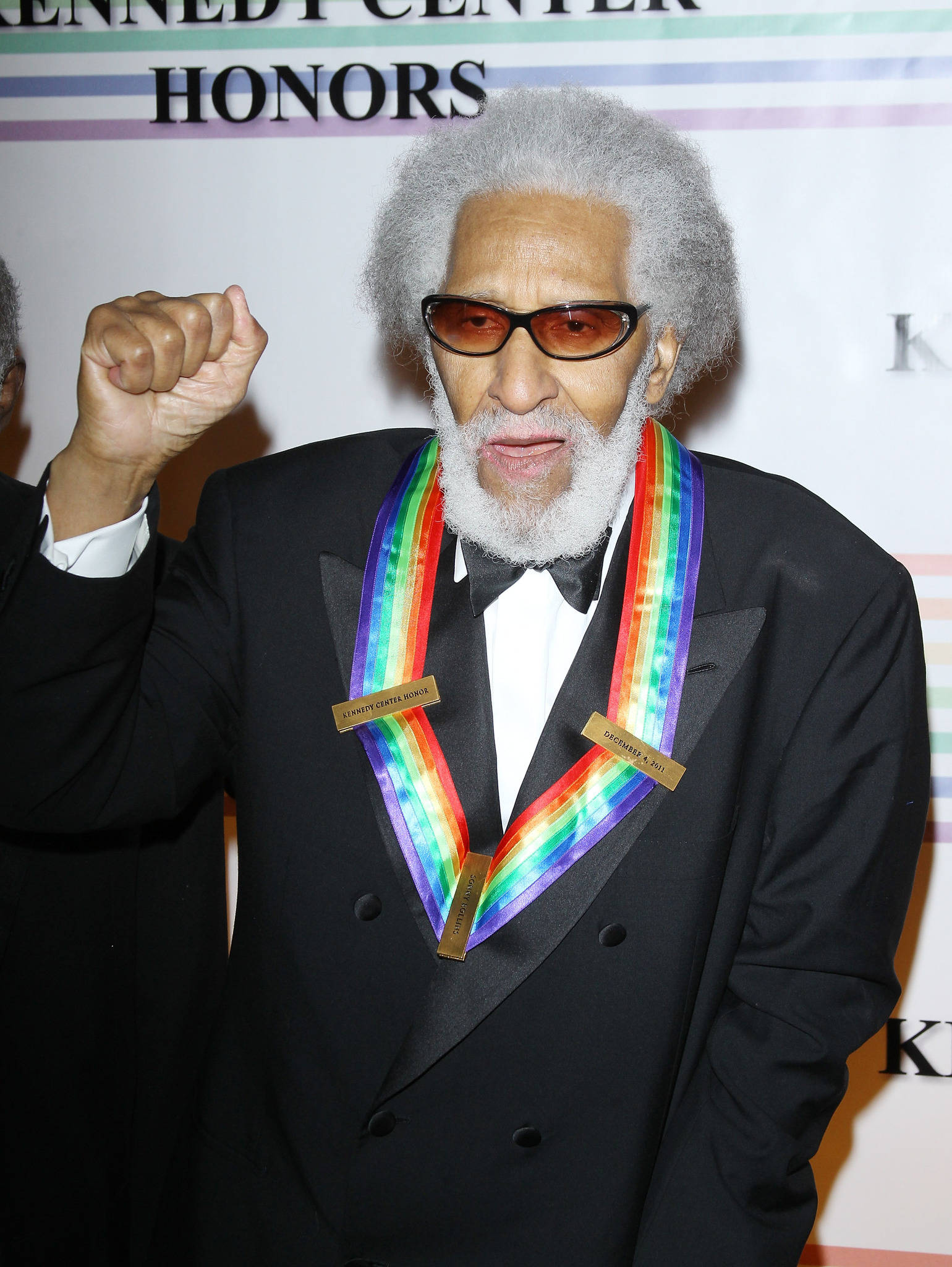 爵士次中音薩克斯風手 Sonny Rollins 壁紙 — 一位灰髮戴太陽眼鏡的長者在甘迺迪中心榮譽獎活動中，佩戴彩虹獎章並舉起拳頭。