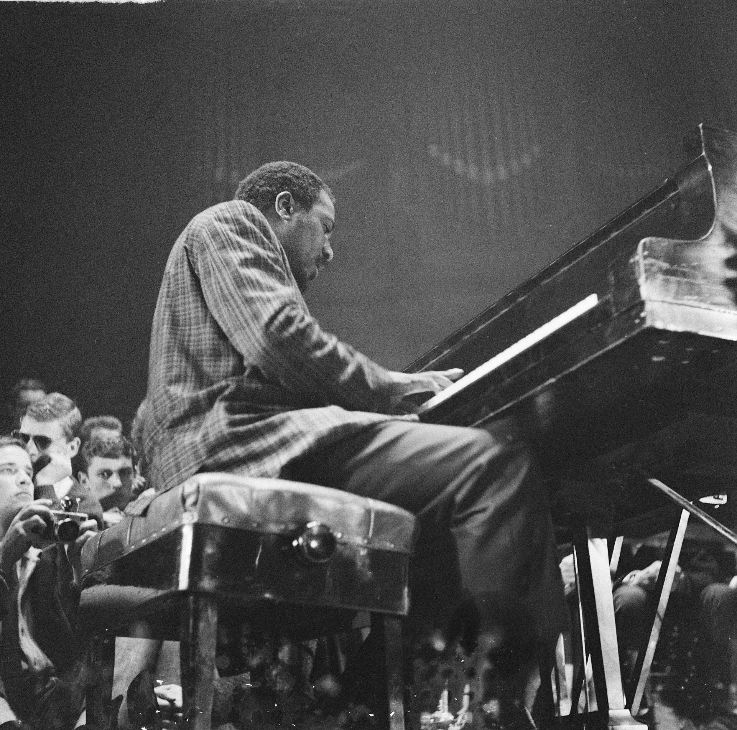 爵士鋼琴大師 Thelonious Monk 演奏古典鋼琴 壁紙 — 一位穿著格仔恤衫的爵士鋼琴家在舞台燈光下彈奏三角鋼琴，觀眾專注地觀看。