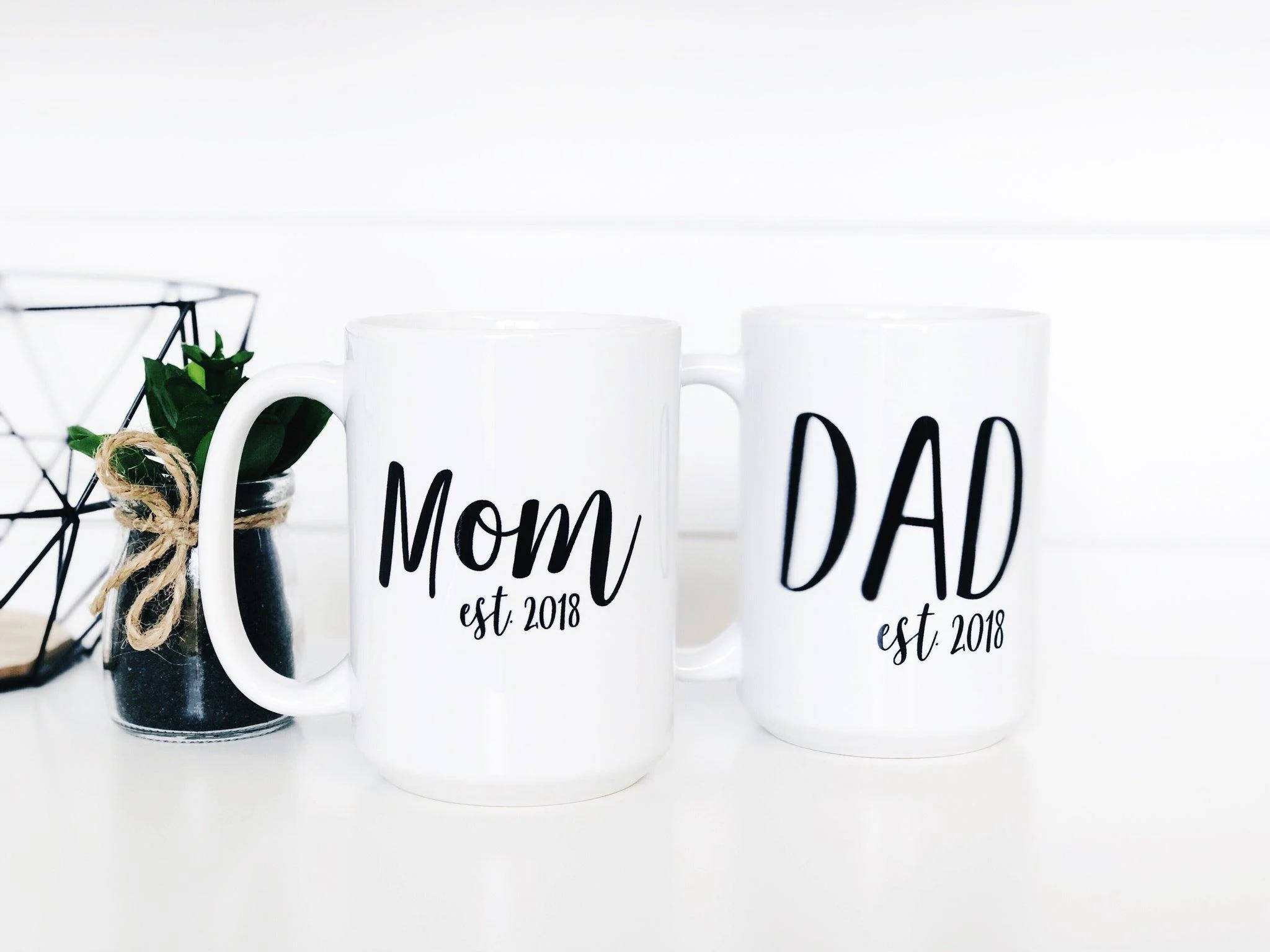 父母杯墊圖案 壁紙 — 兩個白色馬克杯，上面寫著 Mom and Dad est 2018，旁邊放著一個幾何線條鐵籃裡的小盆栽，放在明亮的白色表面上。