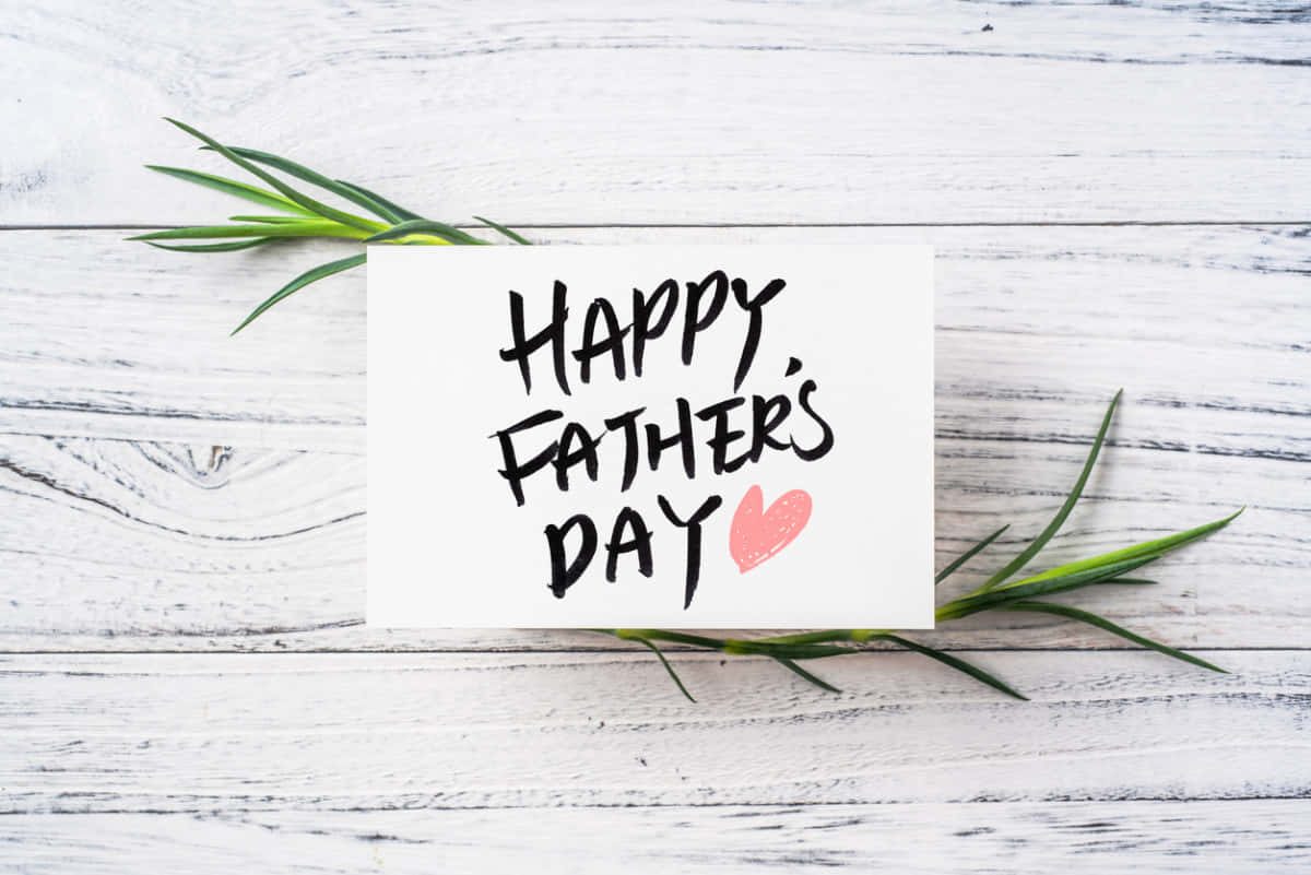 父親節賀卡木質表面圖 壁紙 — 一張白色卡片，上面有手寫的「HAPPY FATHER'S DAY」和一個粉紅色的心形，放在白色木板上，配以綠色的迷迭香枝。