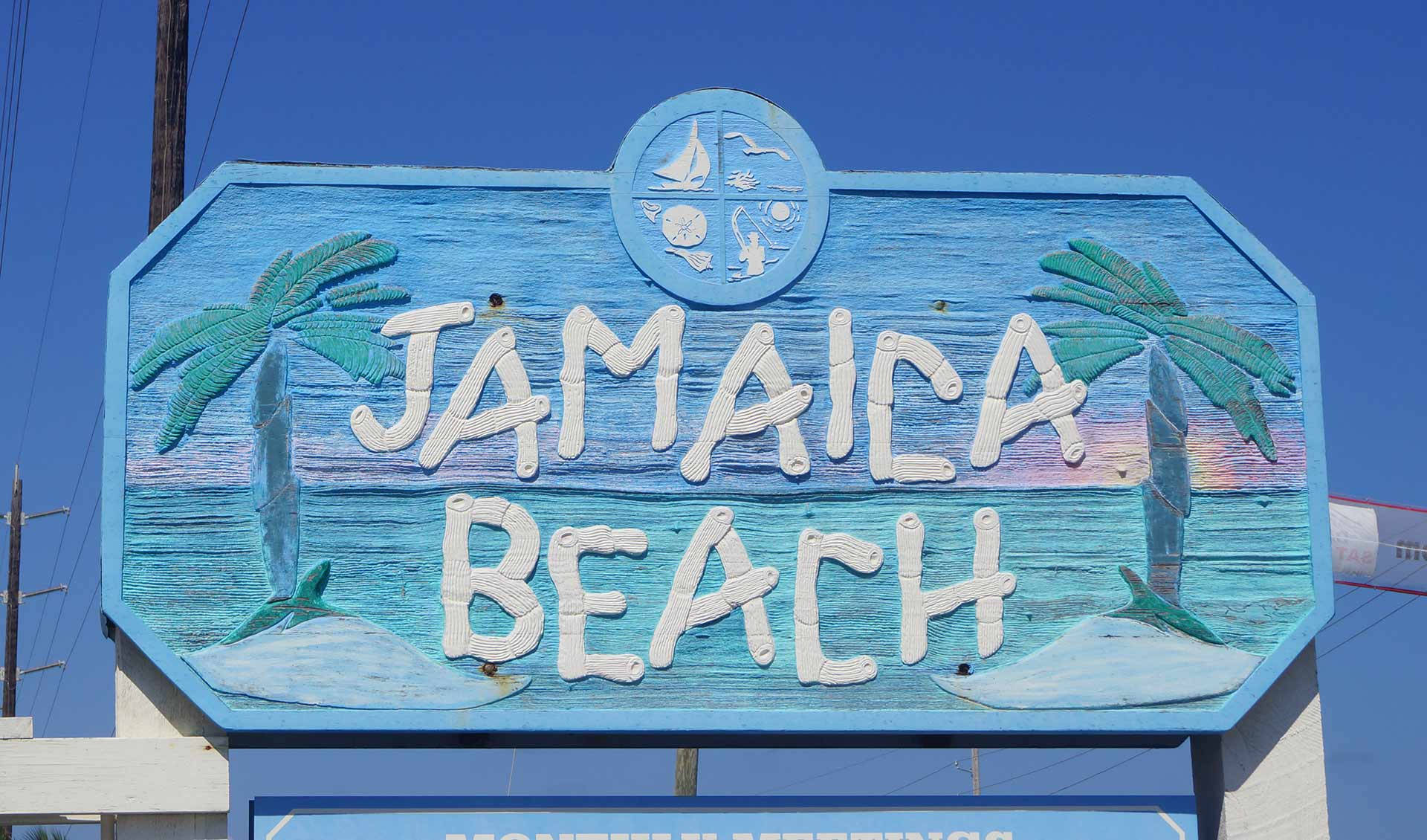 牙買加海灘標誌 壁紙 — 一塊風化的藍色木牌上寫著 Jamaica Beach，上面繪有棕櫚樹和海洋波浪，背景是晴朗的天空。