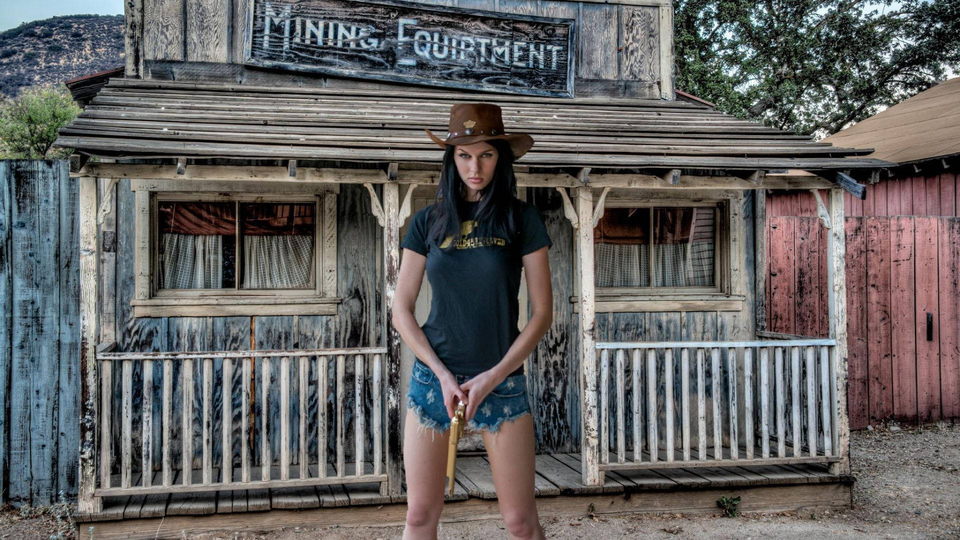 牛仔女持槍 壁紙 — 一位戴著牛仔帽的女性自信地站在一棟寫有「MINING EQUIPMENT」的風化木造建築前，背景是塵土飛揚的西部場景。