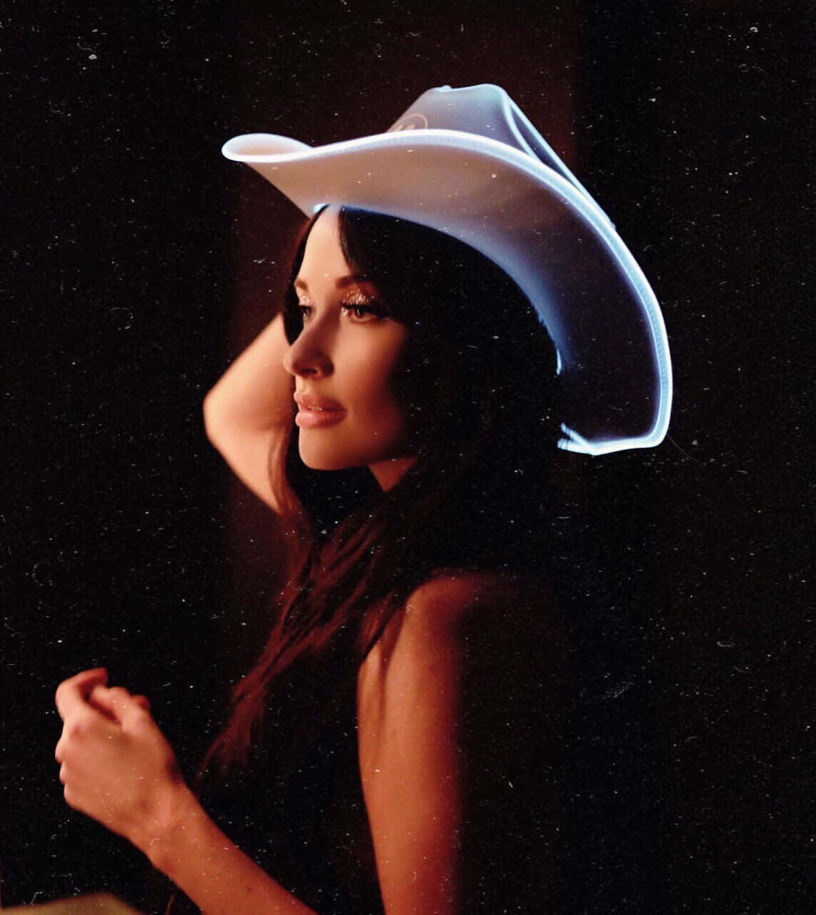 牛仔風美學：音樂人Kacey Musgraves 壁紙 — 一位戴著發光白色牛仔帽的女性，在深色斑點背景前擺姿勢，柔和光線凸顯她的側面輪廓。