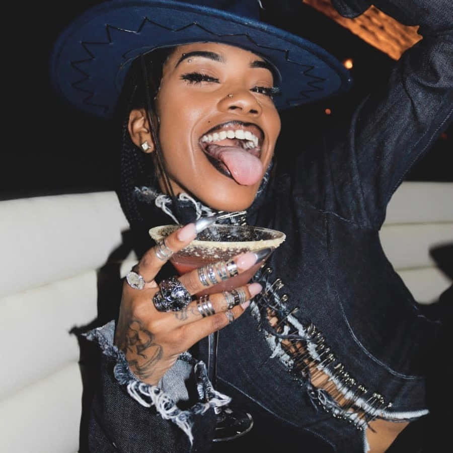牛仔風美學：饒舌歌手Rico Nasty 壁紙 — 一位戴藍色帽子和牛仔外套的快樂女性，在夜晚拿著雞尾酒，頑皮地伸出舌頭。