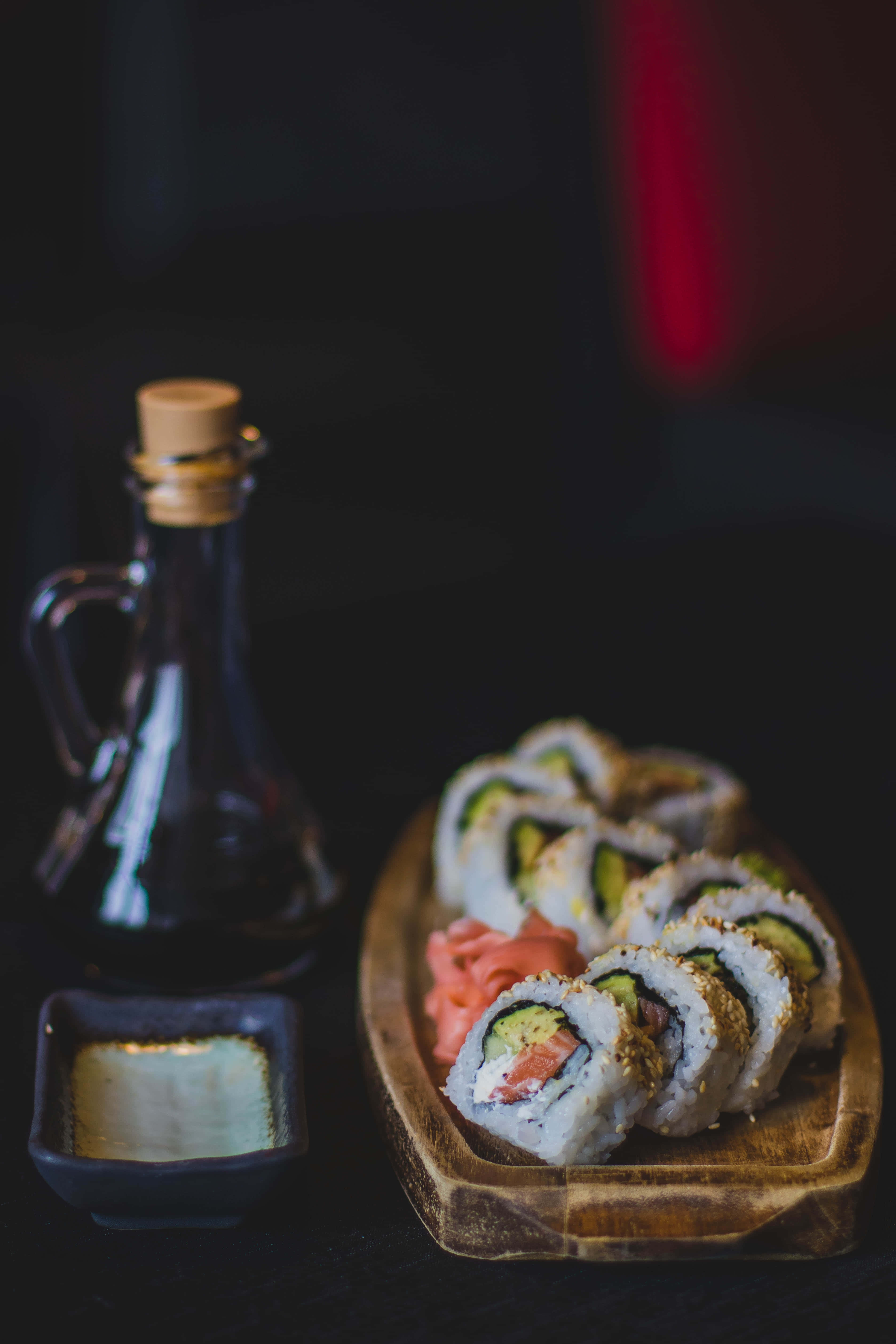 Sushi 桌布 壁紙 — 牛油果三文魚壽司卷放在木盤上，旁邊有醬油和醃薑，環境燈光昏暗。