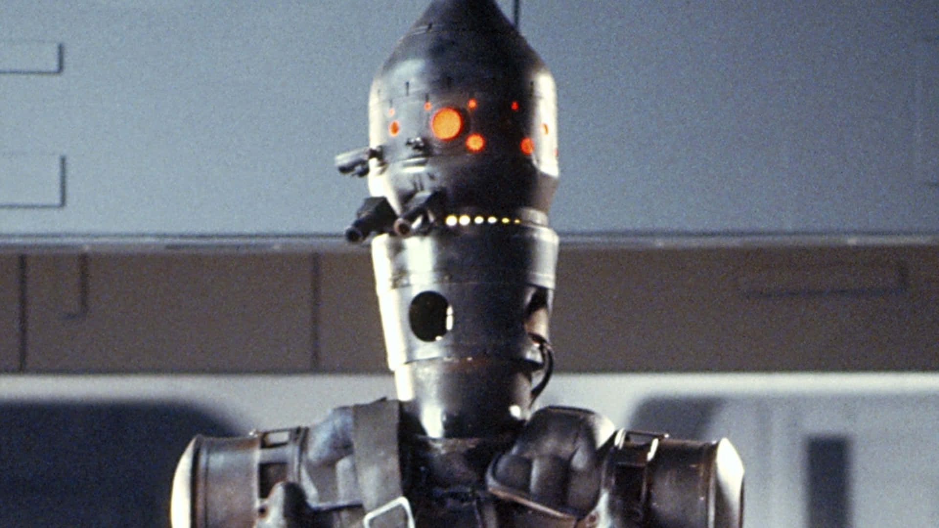 Ig-88 桌布 壁紙 — 特寫一個帶有發光橙色眼睛和機械細節的威脅性黑色機器人，背景是灰色工業牆壁。