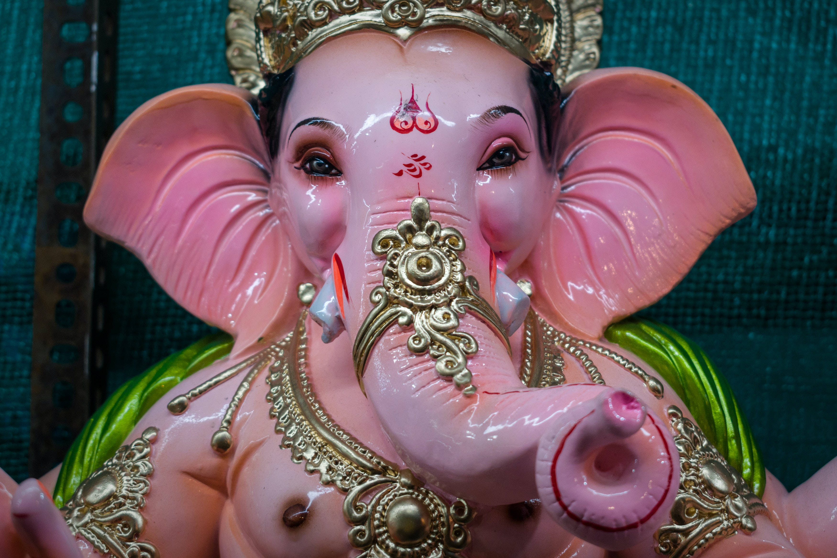 Ganesh 桌布 壁紙 — 特寫一個帶有金色珠寶和紅色標記的光澤粉紅色象頭神雕像，背景為藍綠色。