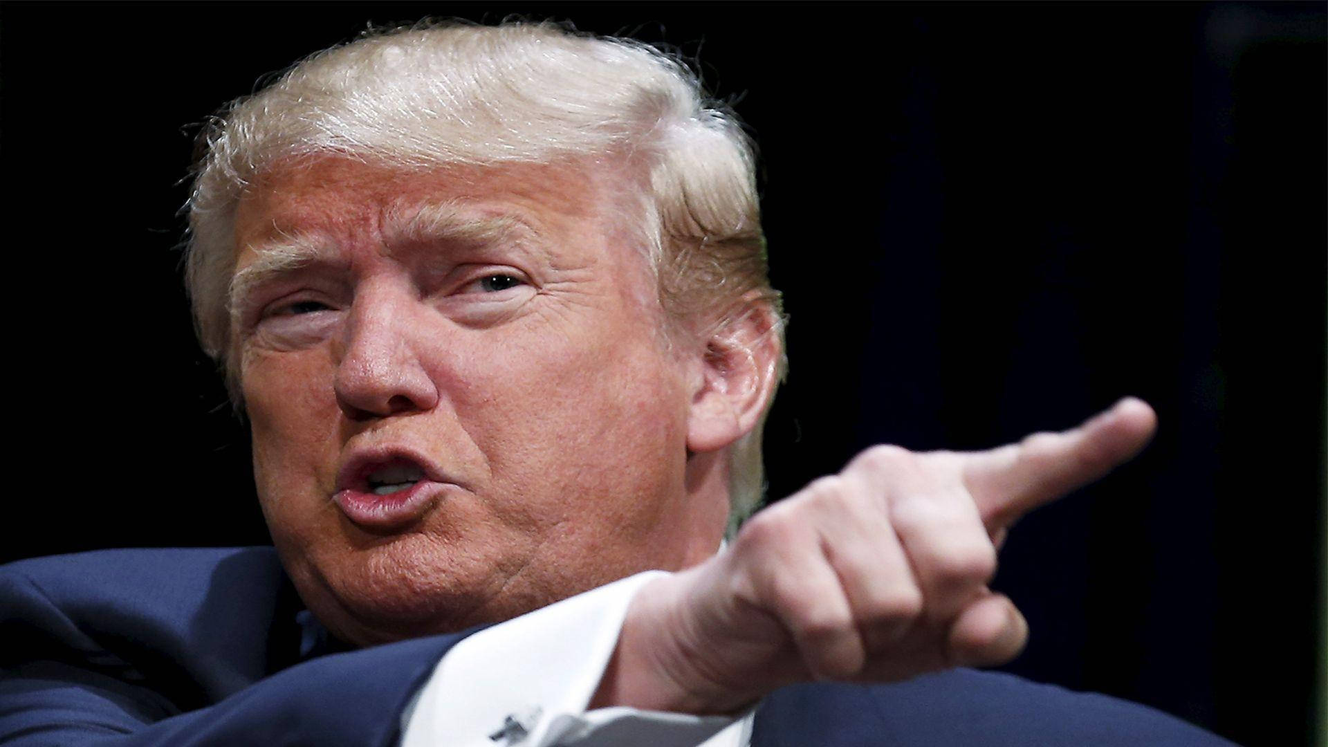 donald trump 桌布 壁紙 — 特寫一名金髮男子身穿西裝，在深色背景下以強烈表情向前指。