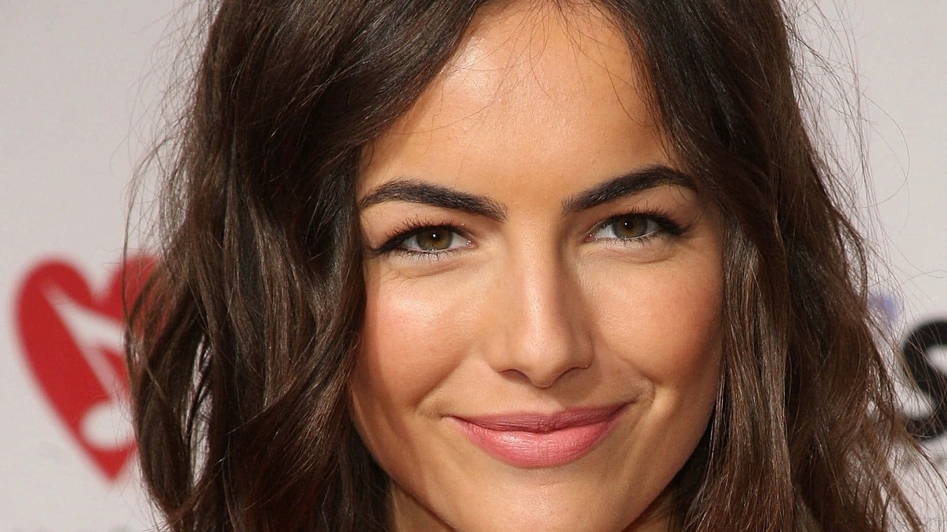 Camilla Belle 桌布 壁紙 — 特寫肖像，一位微笑的女性，深色波浪頭髮，溫暖的棕色眼睛，背景是模糊的活動場景。