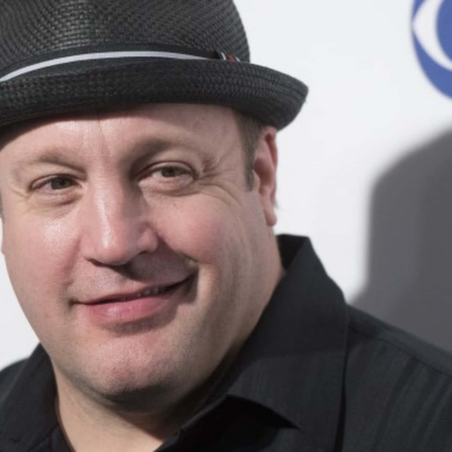 Kevin James 桌布 壁紙 — 特寫肖像，一位微笑的男子戴著黑色軟呢帽和深色襯衫，背景為明亮的白色。