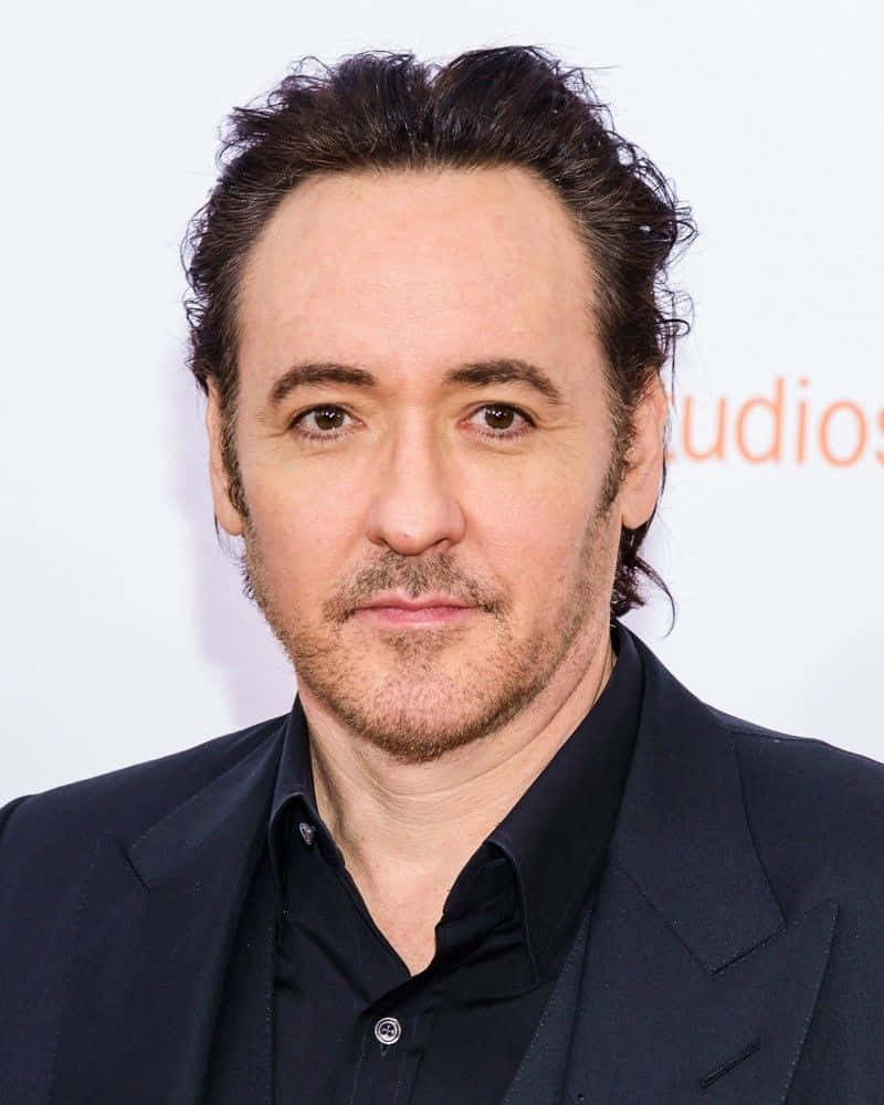 John Cusack 桌布 壁紙 — 特寫肖像，一名深色頭髮、帶鬍渣的男子，身穿黑色西裝和襯衫，背景為白色。