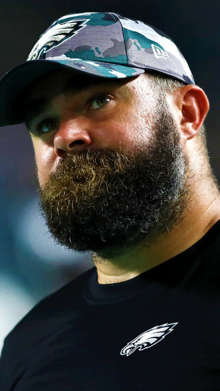 Jason Kelce 桌布 壁紙 — 特寫肖像，一名留鬍鬚的男子在球場燈光下戴著費城老鷹隊迷彩帽和穿著球隊襯衫。