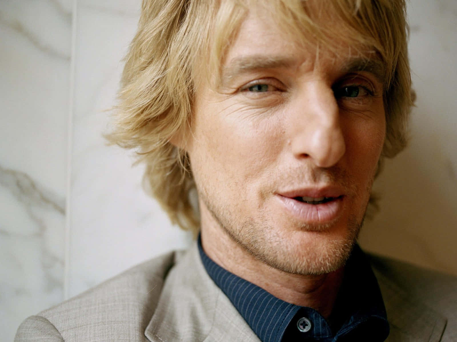 Owen Wilson 桌布 壁紙 — 特寫肖像：一名金髮男子，帶有輕微鬍渣，身穿米色西裝和藍色襯衫，背景為大理石牆。