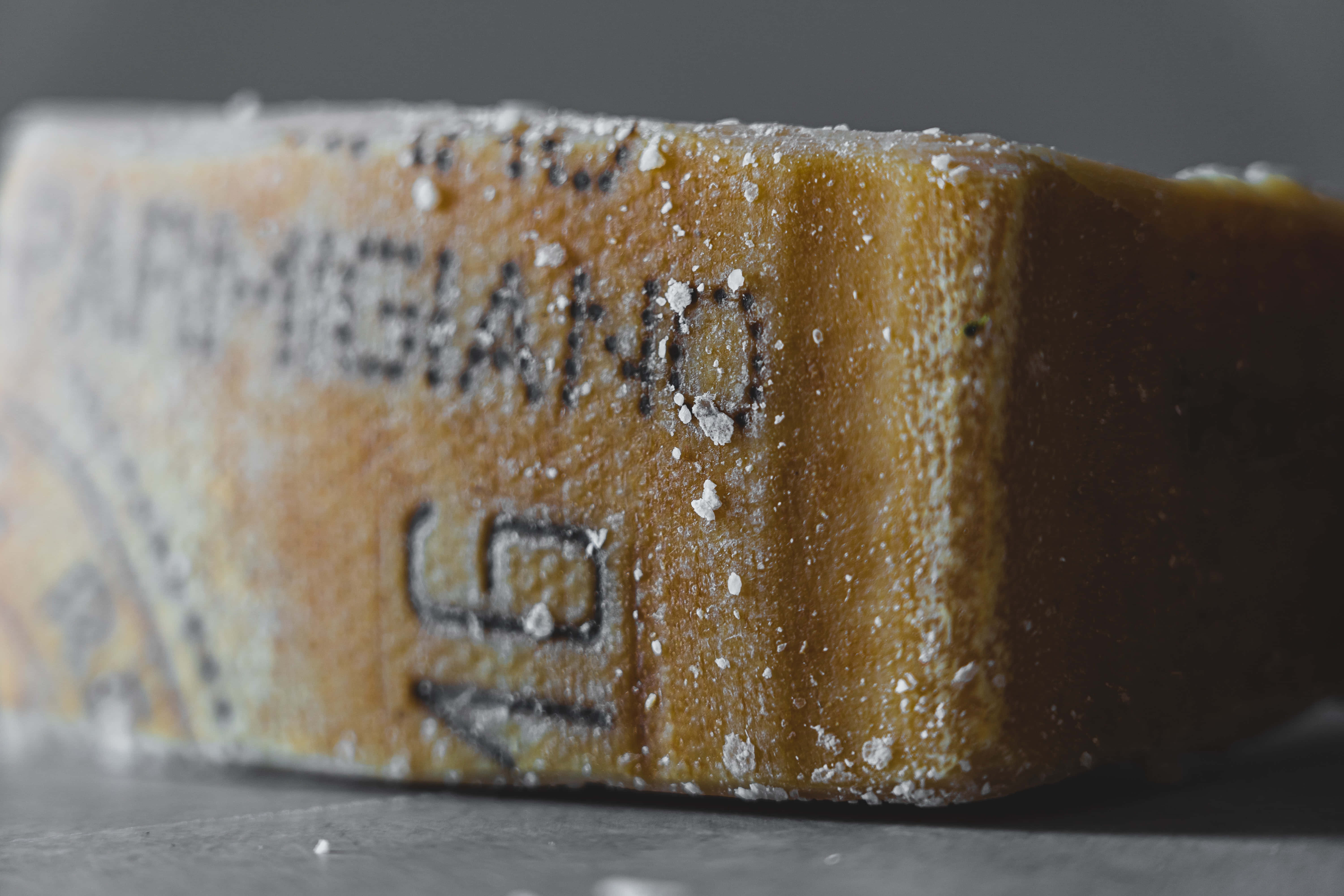 Cheese 桌布 壁紙 — 特寫鏡頭：一塊鄉村風格的帕瑪森雷吉亞諾芝士楔形塊，撒上細白晶體，置於深灰色背景上。