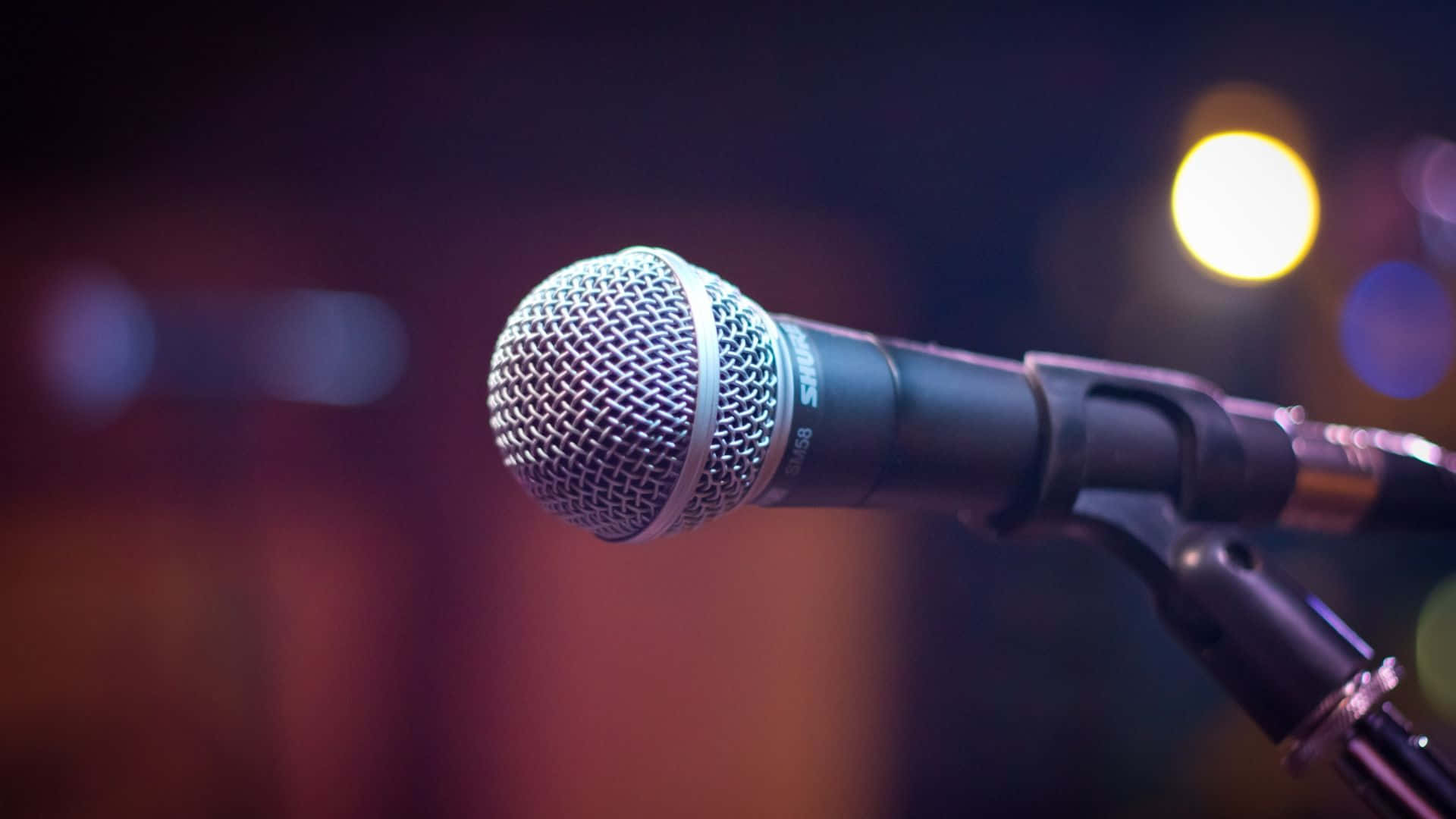 Microphone 桌布 壁紙 — 特寫鏡頭：一支Shure咪高峰放在咪架上，處於溫暖舞台燈光下，背景模糊。