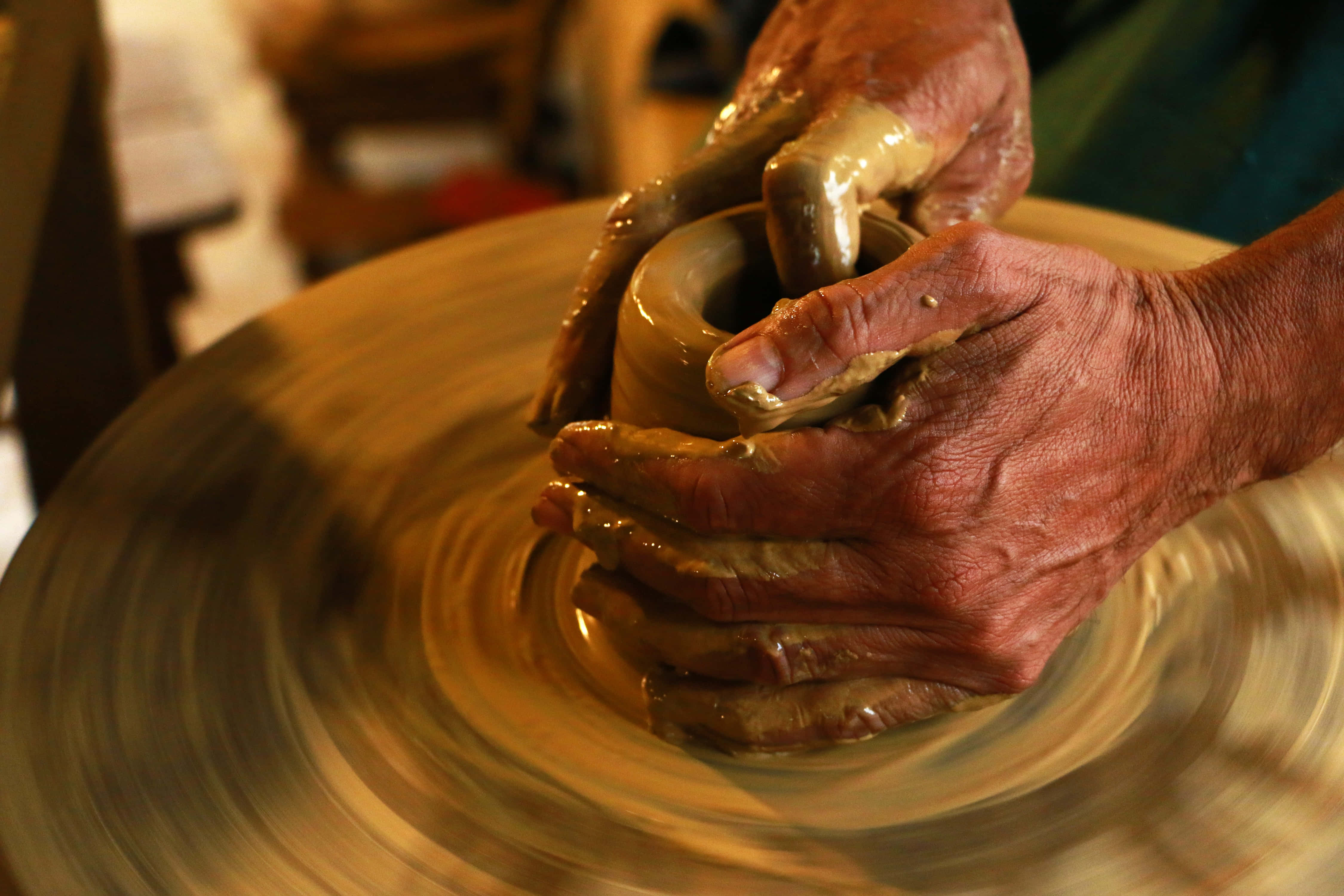 hands 桌布 壁紙 — 特寫鏡頭，一雙歷經風霜的手在溫暖的金色光線下，熟練地在旋轉的陶輪上塑造濕粘土。