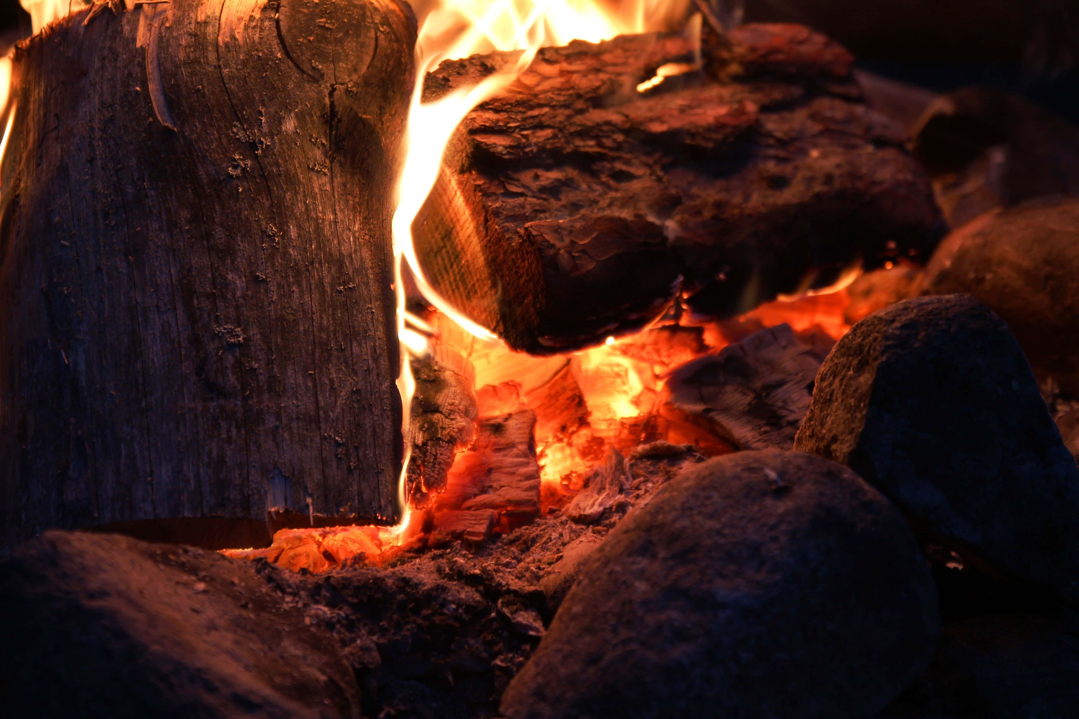 Campfire 桌布 壁紙 — 特寫鏡頭：在一個被燒焦餘燼和石頭包圍的黑暗營火坑中，噼啪作響的木柴閃爍著橙色火焰。