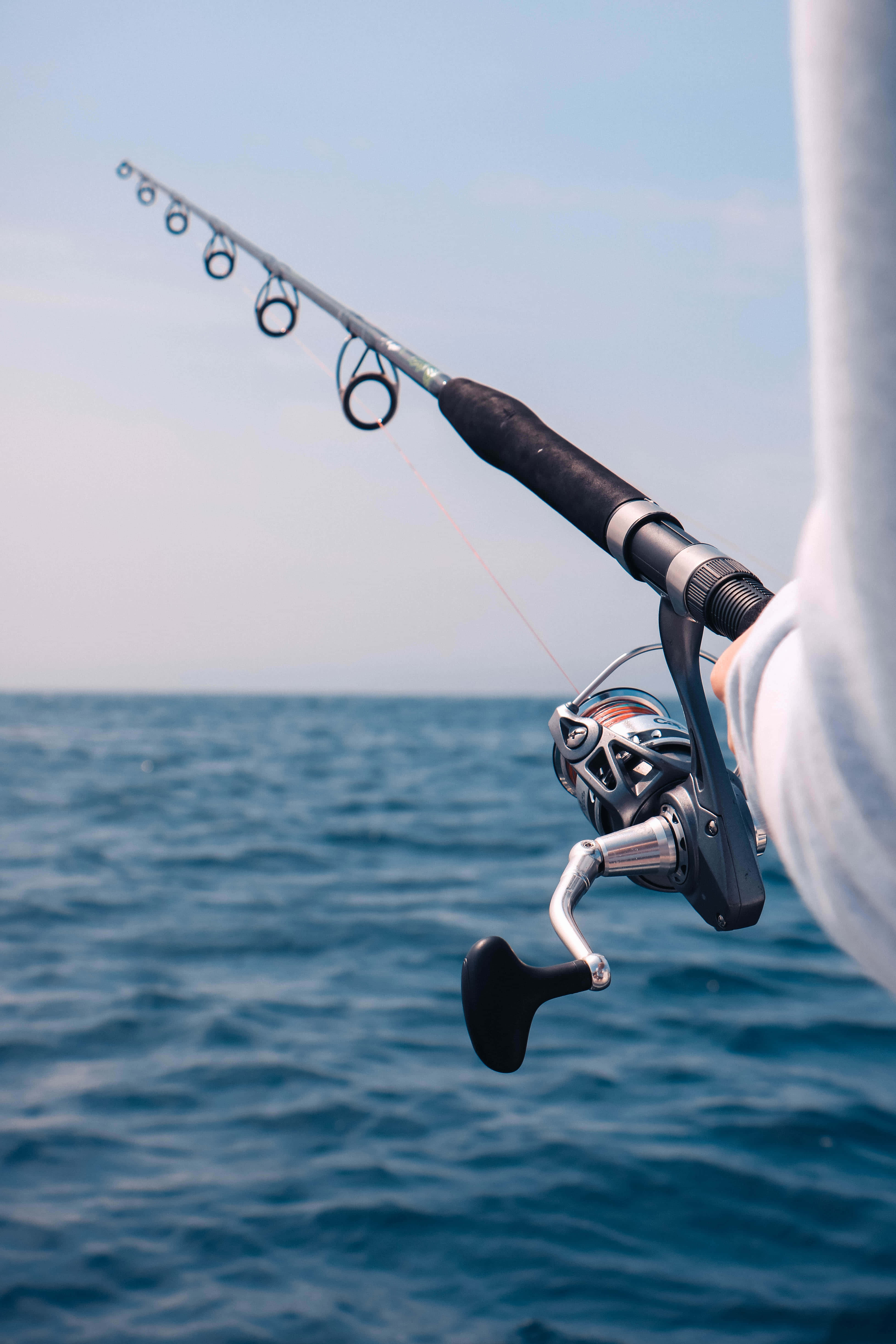 Fishing Rod 桌布 壁紙 — 特寫鏡頭：晴朗天空下，有人手持魚竿和捲線器，向開闊海洋拋投。