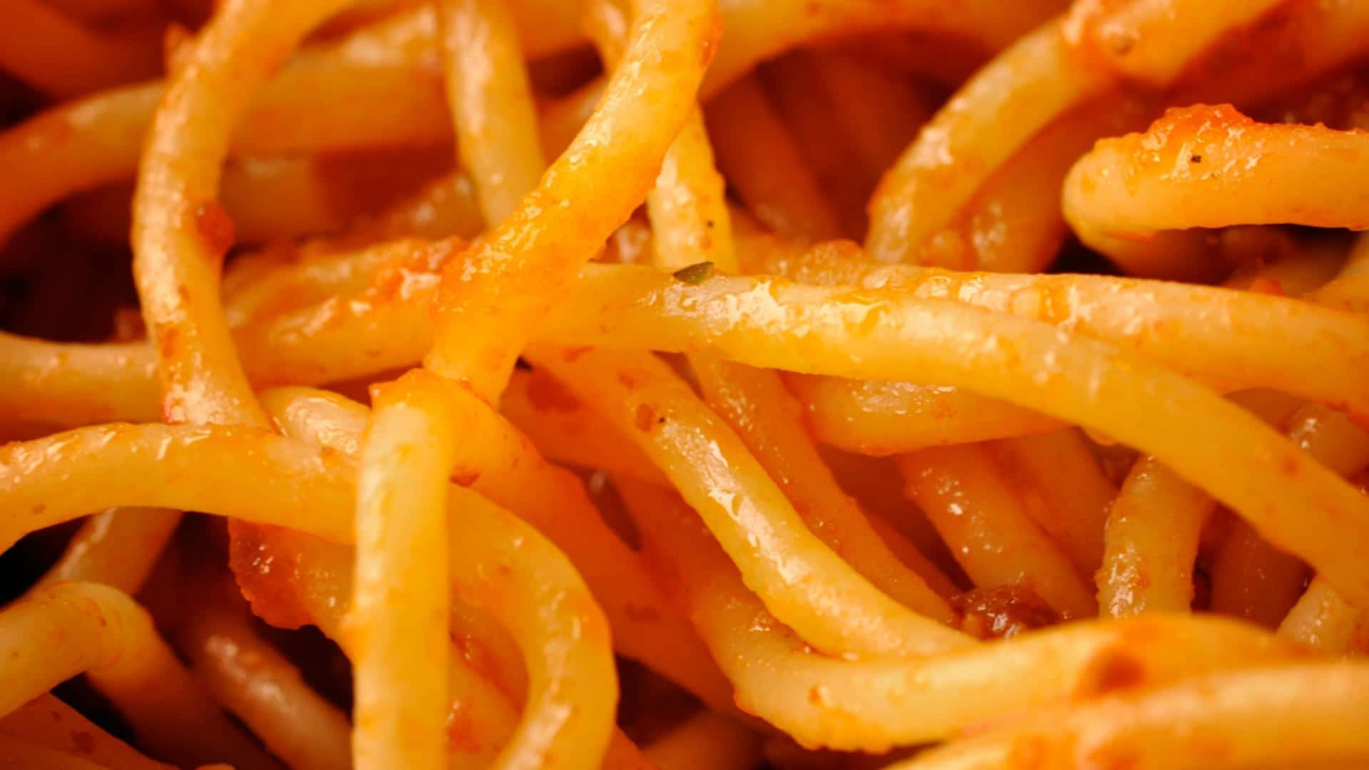 Pasta 桌布 壁紙 — 特寫鏡頭：濃郁番茄醬包裹的亮澤意大利麵，可見香草和油在溫暖燈光下閃爍。