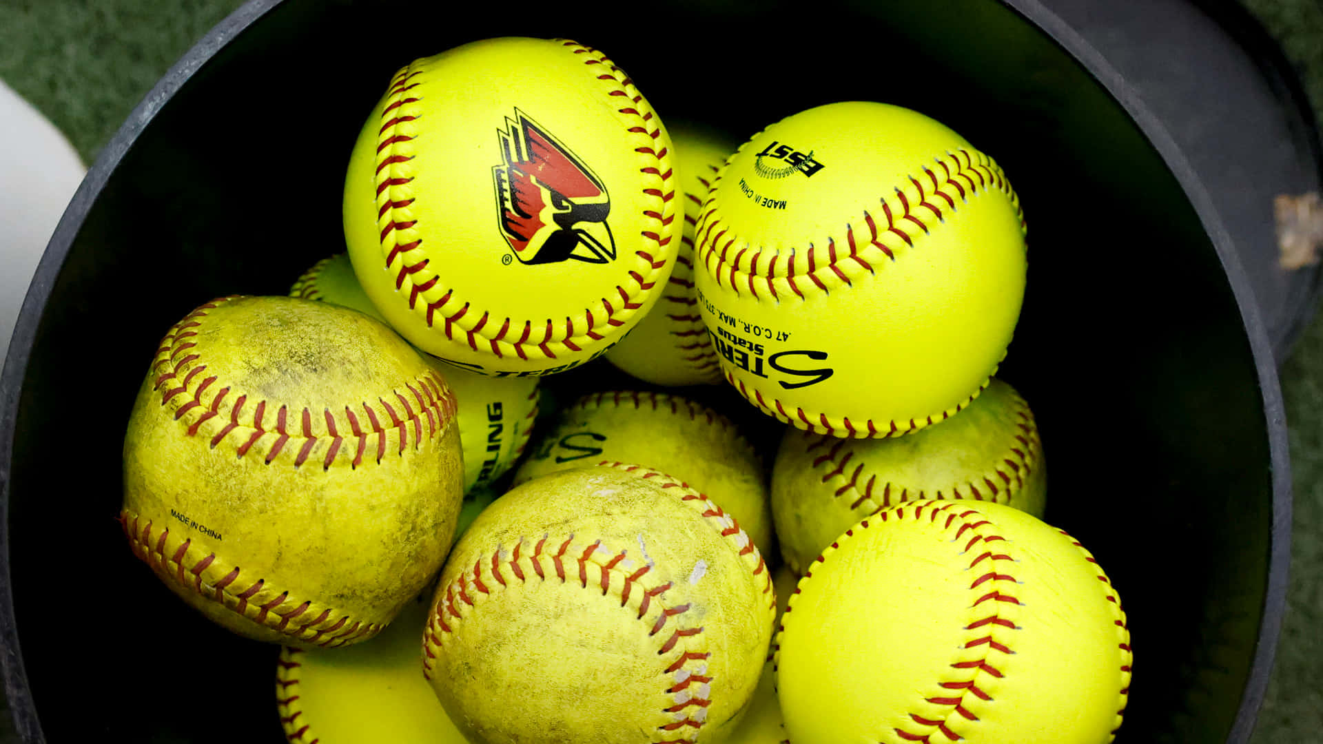 Softball 桌布 壁紙 — 特寫鏡頭：綠色草地場上黑色桶內堆疊的亮黃色壘球，帶紅色縫線。